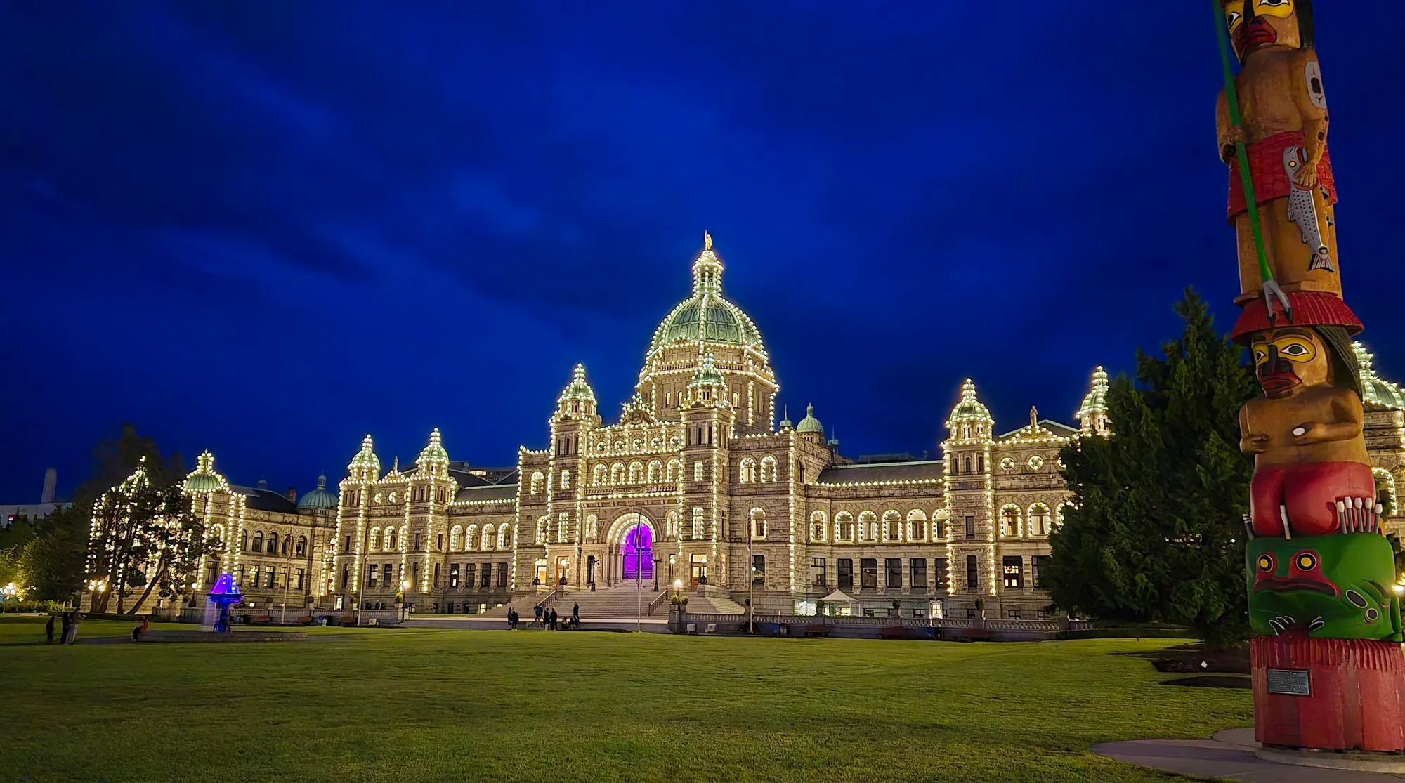 The British Columbia Parliament Buildings lit at night, a totem pole in the foreground