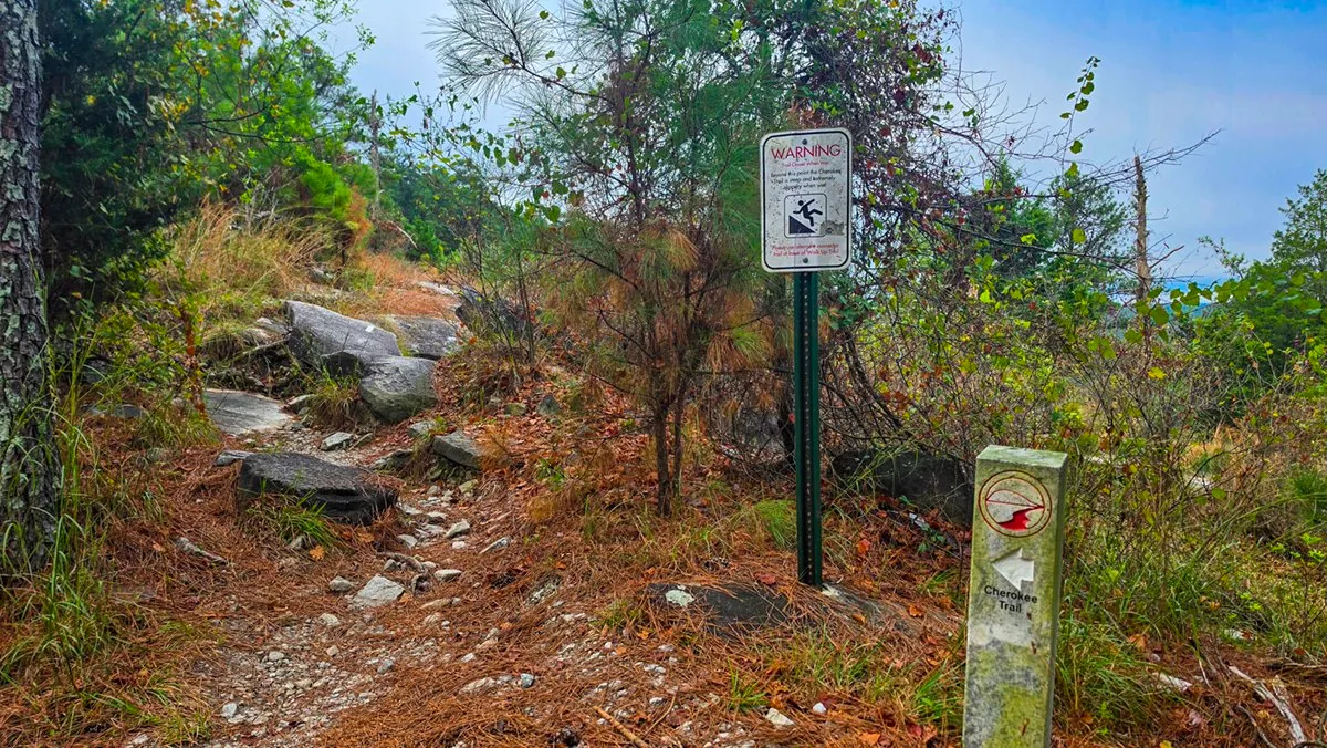 A red and white warning sign on a post beside a Cherokee Trail marker, at the edge of rocky ground and pines