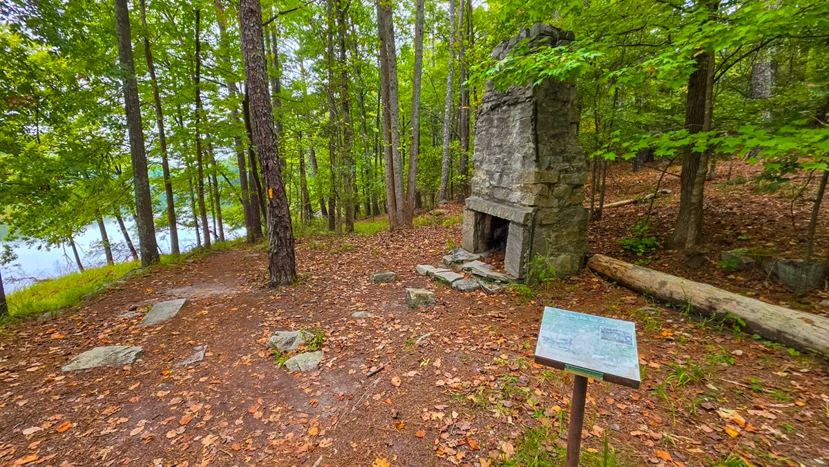 An old stone chimney standing alone in the woods by the lake, an interpretive sign in front of it
