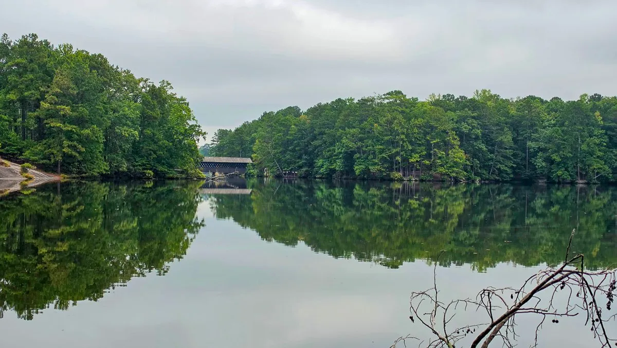 The covered bridge seen far across calm Stone Mountain Lake, between wooded shores