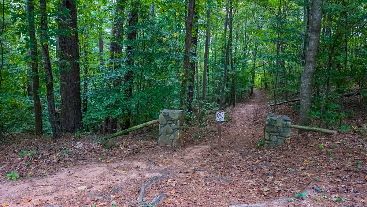 The trail leaving the lot between two stone pillars, a No Bicycles sign on a post between them