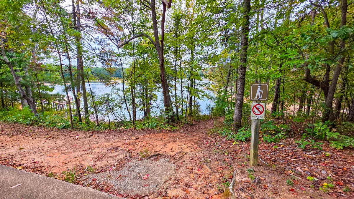 A trail post with a hiker sign and a No Bicycles sign where the path leaves the lot, the lake below through the trees