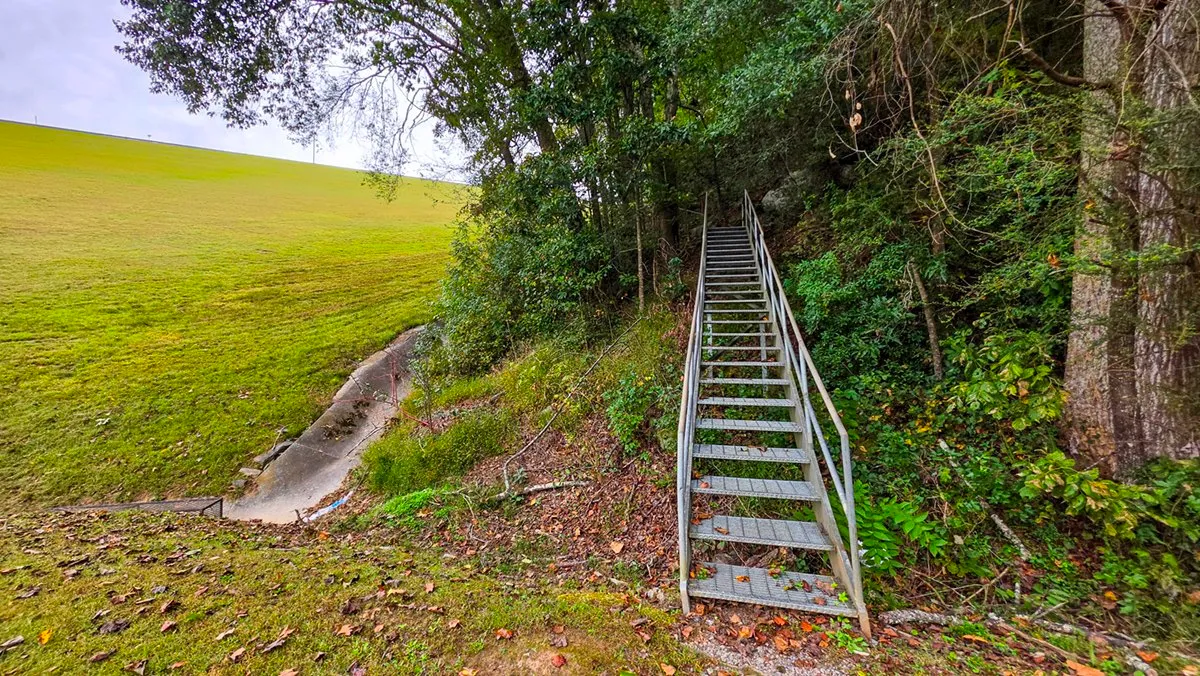 A steep metal staircase climbing the wooded bank beside the grassy face of the dam