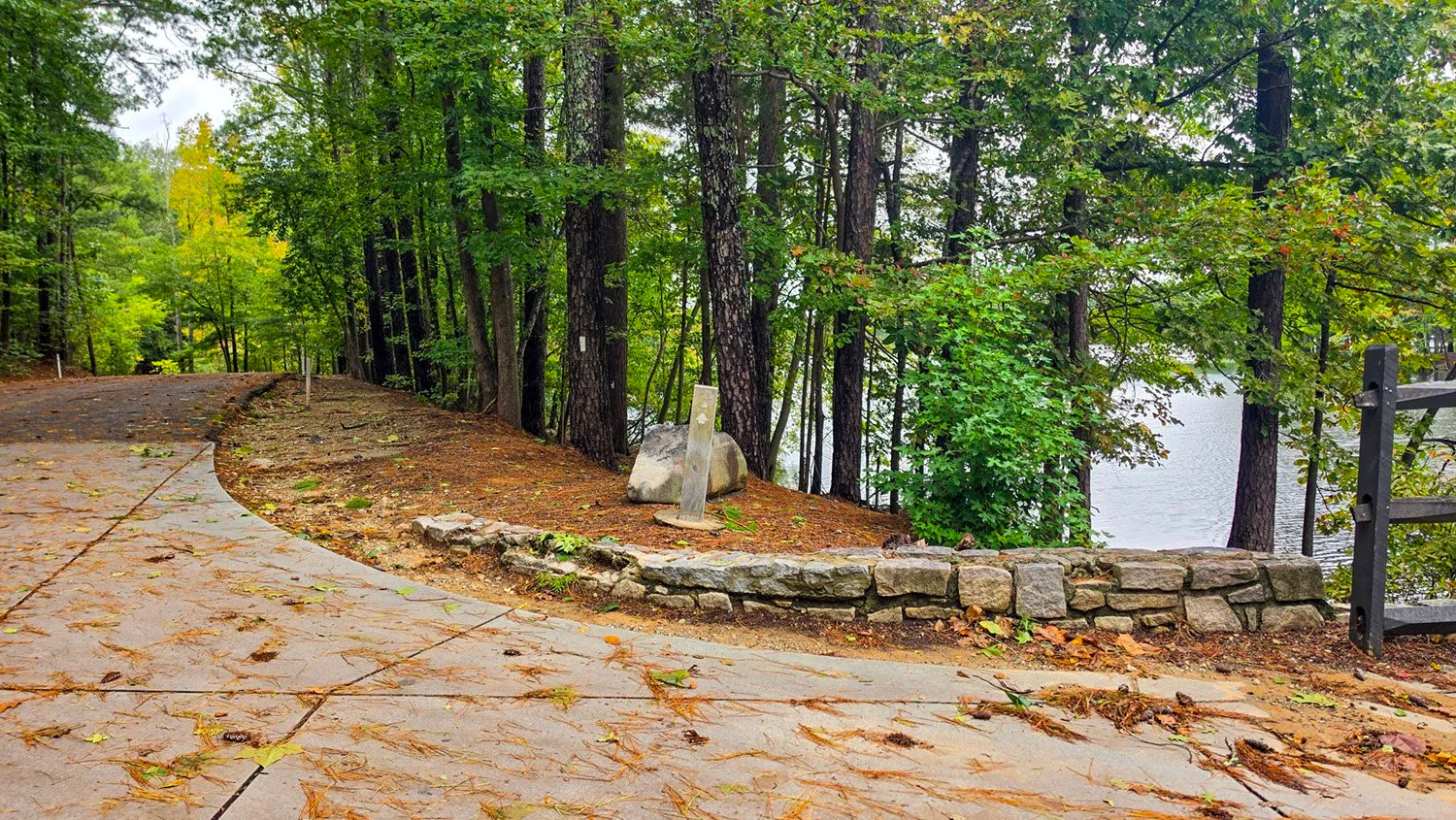 A paved path curving past a low stone wall at the lake's edge, a trail post in front of a boulder in the pine needles and tall pines over the water
