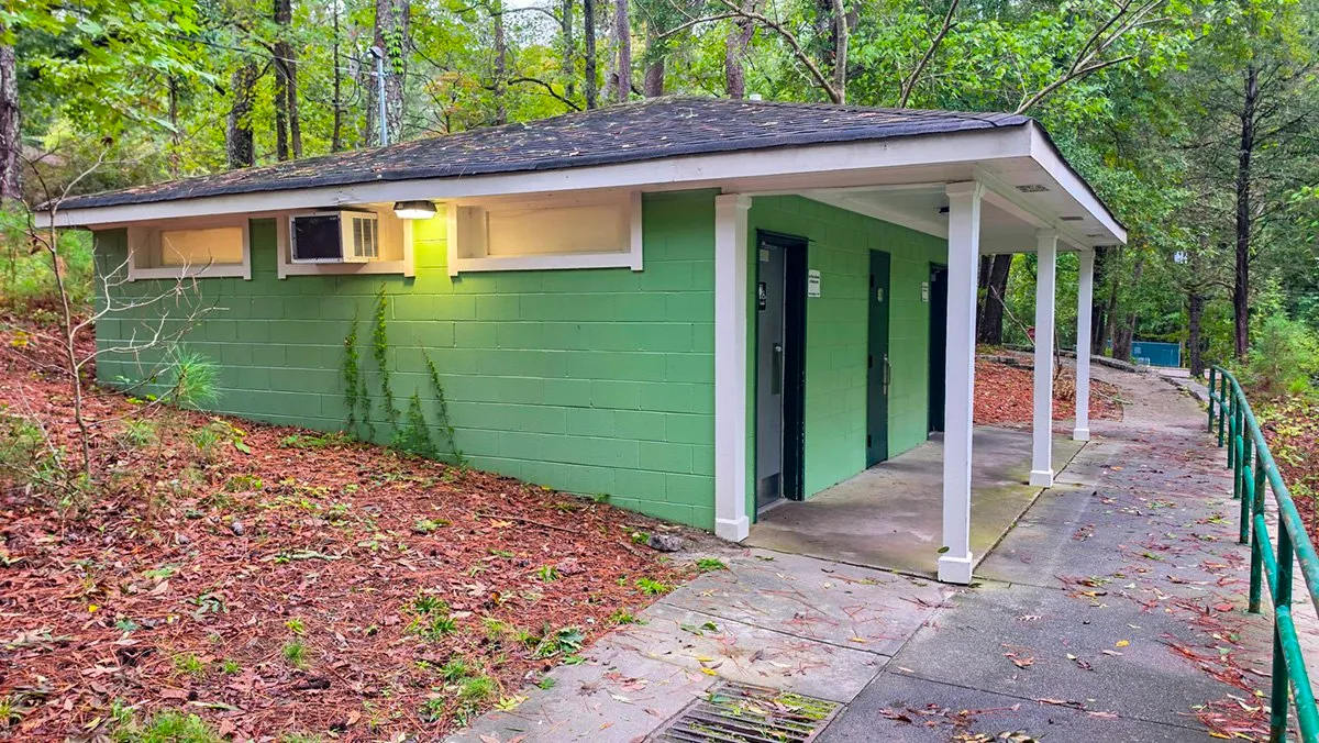 A green block restroom building with white trim and a covered walkway, a railed paved path running past it into the woods