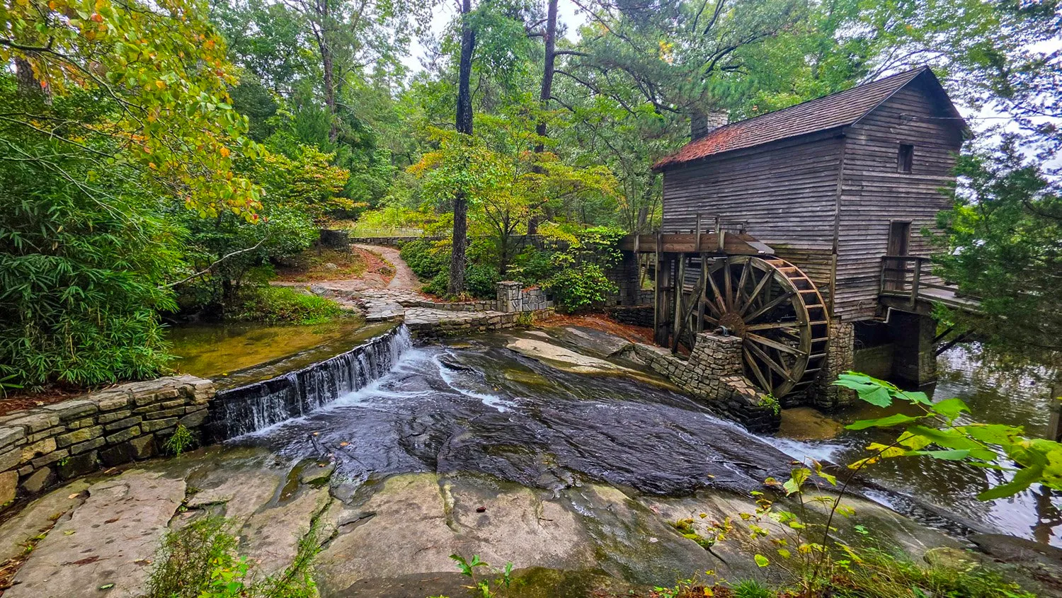 The gray wooden Grist Mill with its large waterwheel, water spilling over a stone spillway and across dark granite below it, green woods around