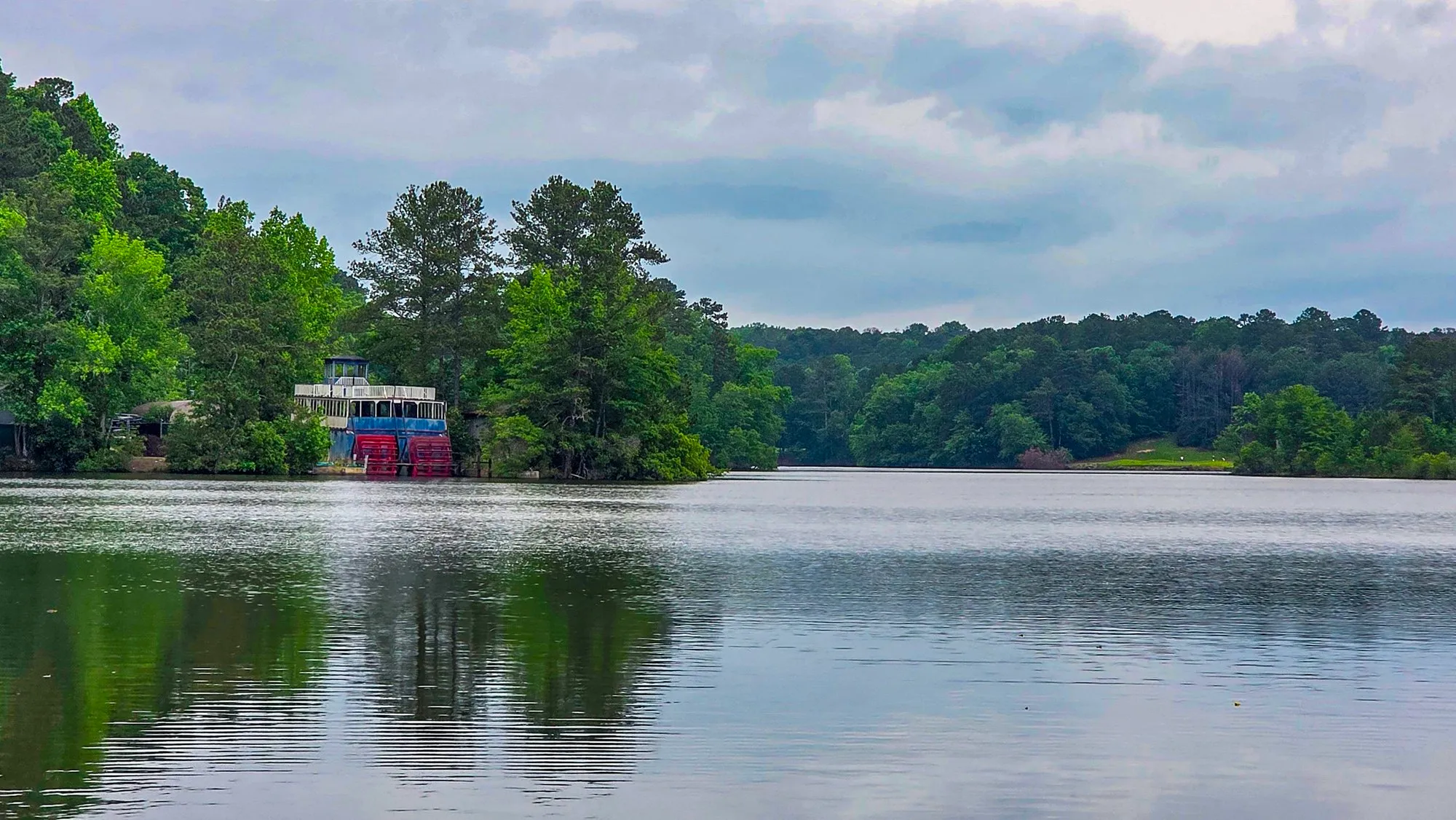 The riverboat seen across the water from the far shore, its blue hull and red wheel at the foot of the trees, the sky gray over the lake
