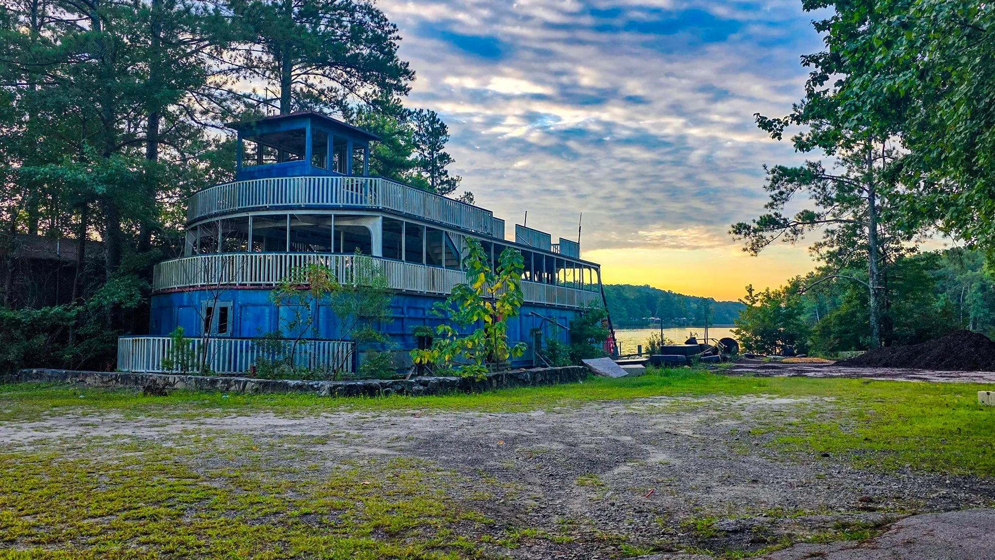 The riverboat up close on its pad, two decks of white railing and the pilothouse, the far shore lit orange behind it and saplings growing against the hull