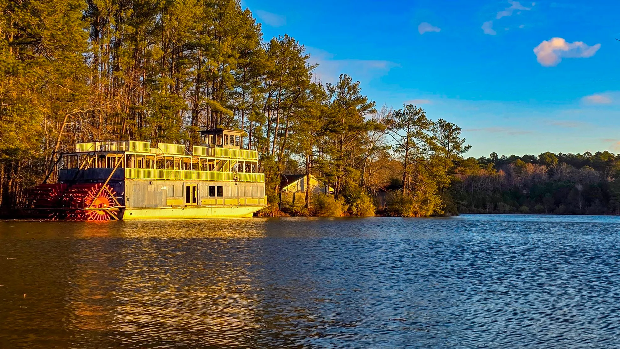 The riverboat afloat on the lake, off its pad, the red stern wheel and the two decks lit gold by the low sun under a blue sky