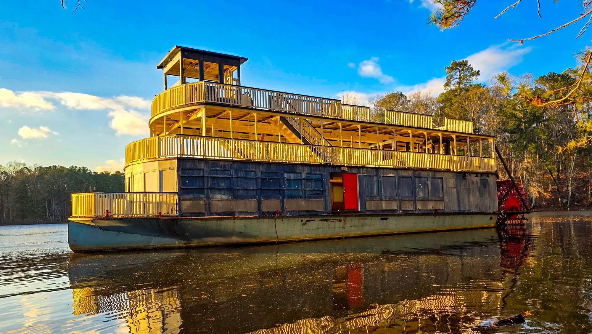 The riverboat adrift from the other side, the stairway between its decks, a red door on the lower deck and the wheel at the far end, the water flat as glass
