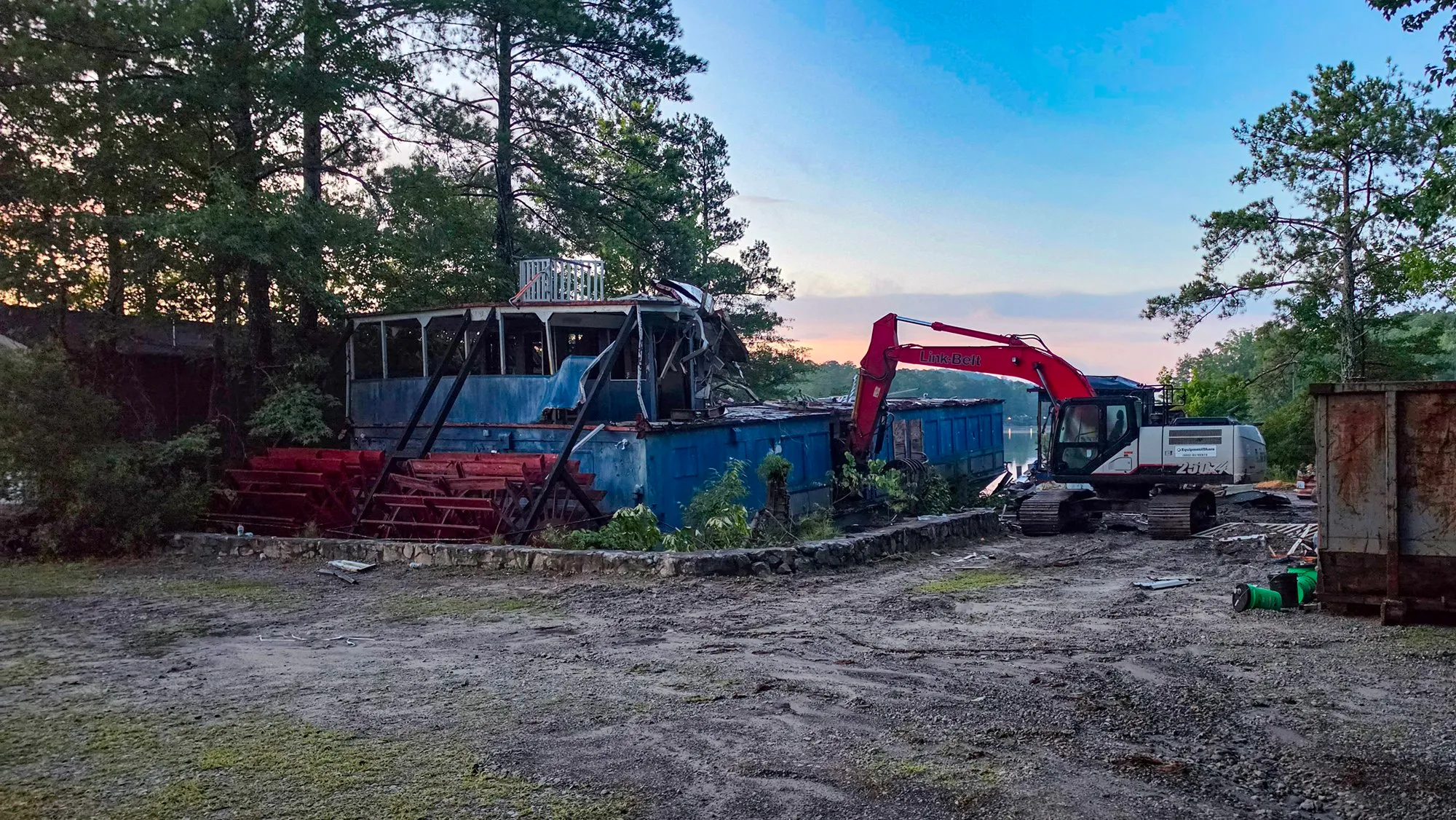 An excavator on the pad with its arm in the riverboat's upper deck, the wheel frames torn off and lying in front of the hull, before sunrise
