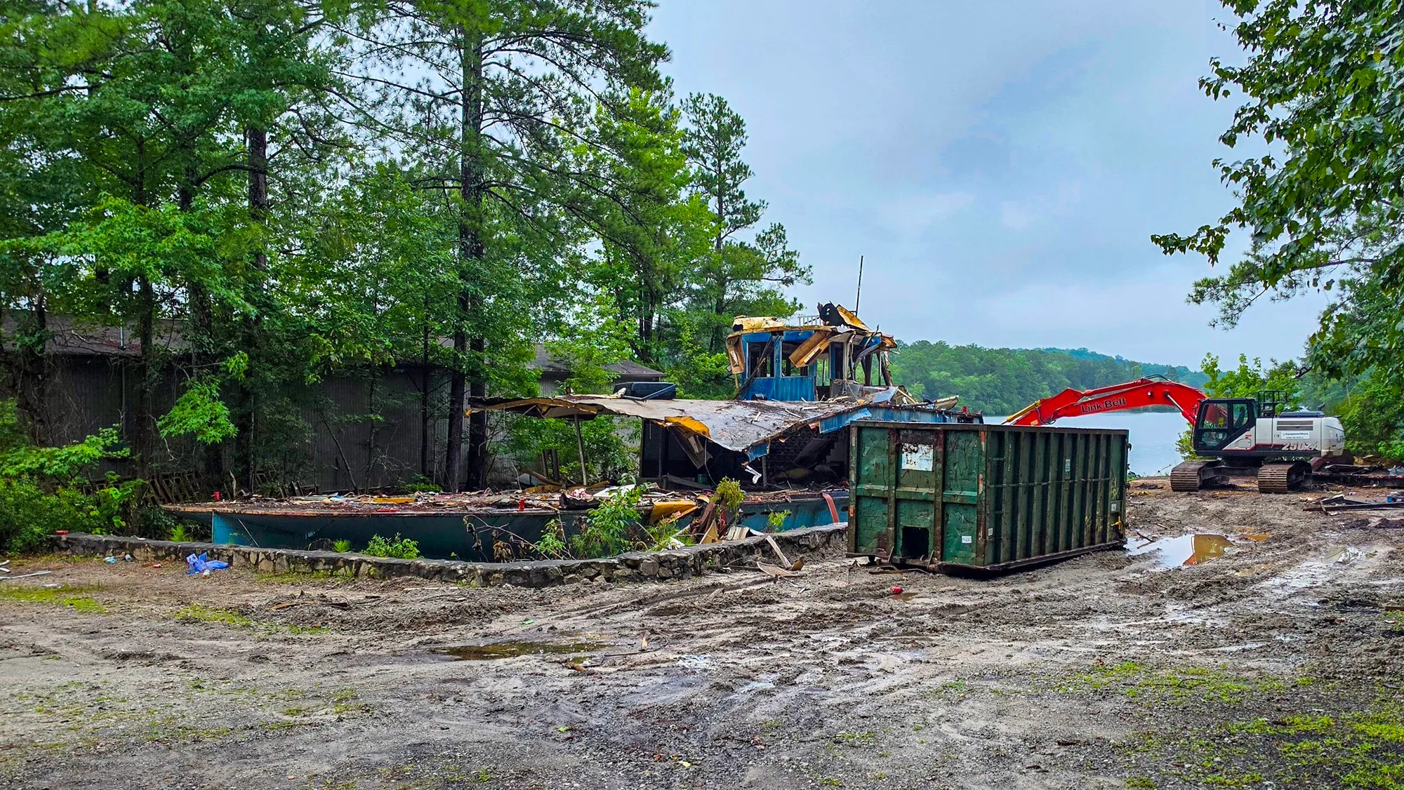 The riverboat's hull folded in on itself on the pad, the pilothouse hanging off the wreck, a green dumpster and the excavator beside it under a gray sky