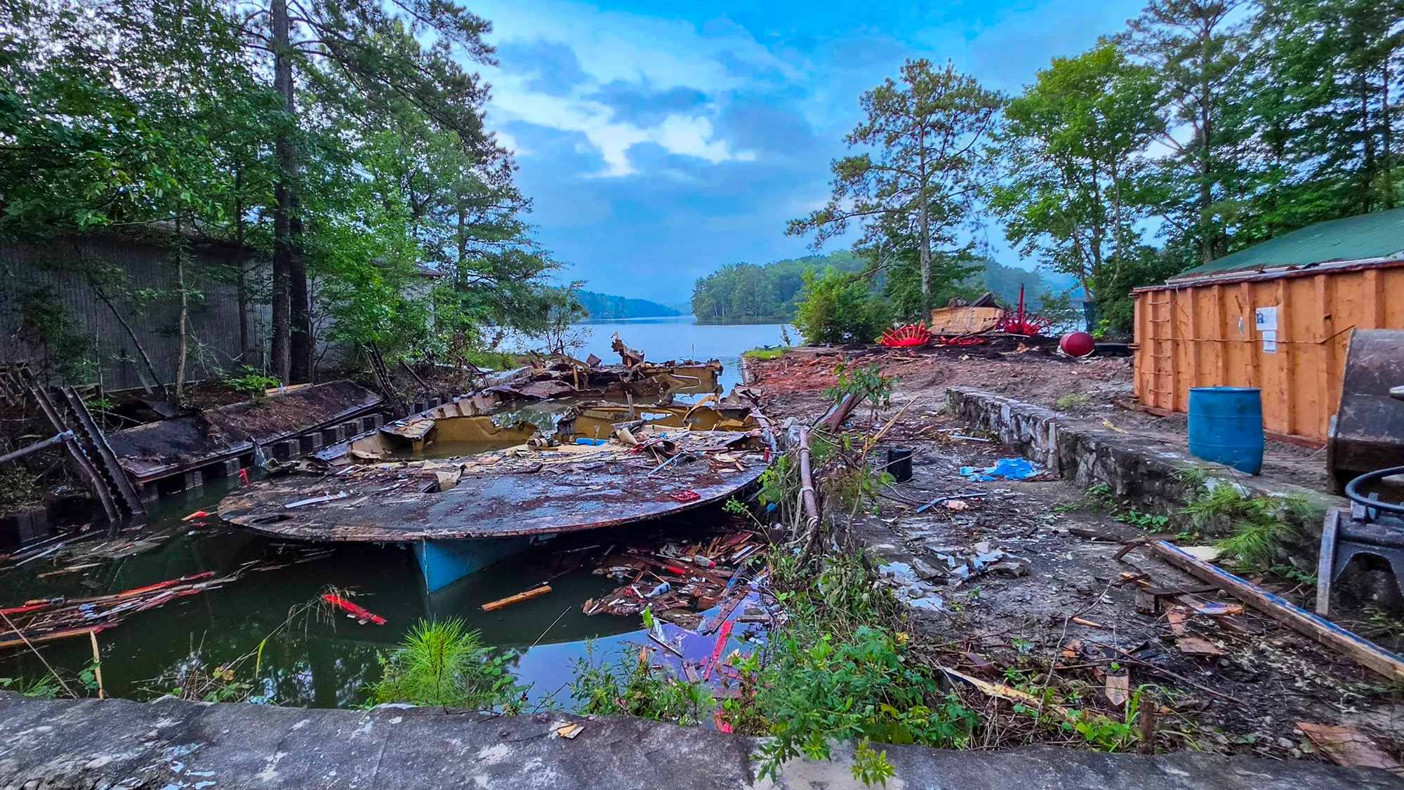 A shell of the riverboat's hull in the water at the seawall, wreckage floating around it, the red wheel frames in a heap on the shore and a new wooden shed going up on the right