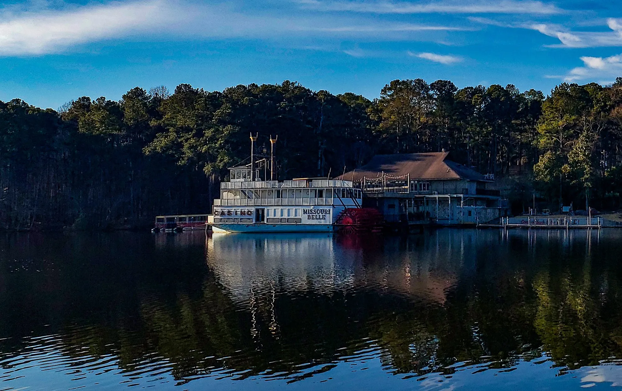 The riverboat dressed as the Missouri Belle at the boathouse pier: CASINO lettered along the lower deck, MISSOURI BELLE beside the red stern wheel, two stacks behind the pilothouse, the white boat doubled in the still lake under a blue sky