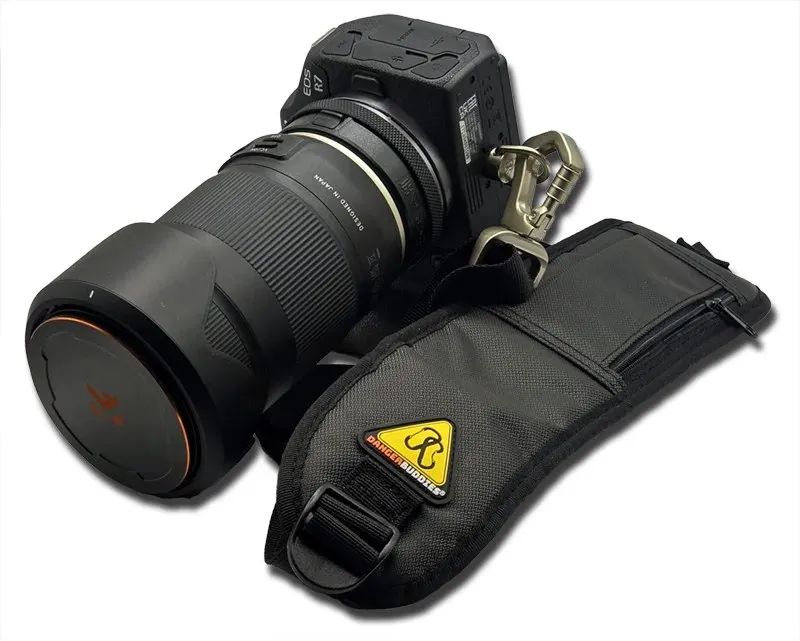 The Danger Buddies S-Curve sling strap beside the R7 and the Tamron, the yellow warning triangle stitched to the black padding and the swivel clip open above it