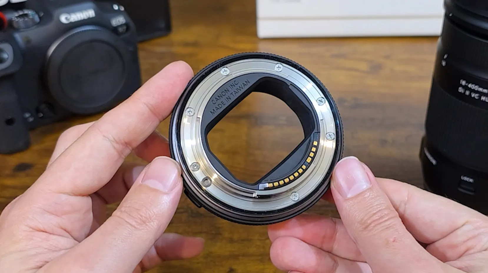 The Canon EF-EOS R mount adapter held in both hands, CANON INC. MADE IN TAIWAN engraved around the opening and the gold contacts along one side, the R7 body and the Tamron on the table behind