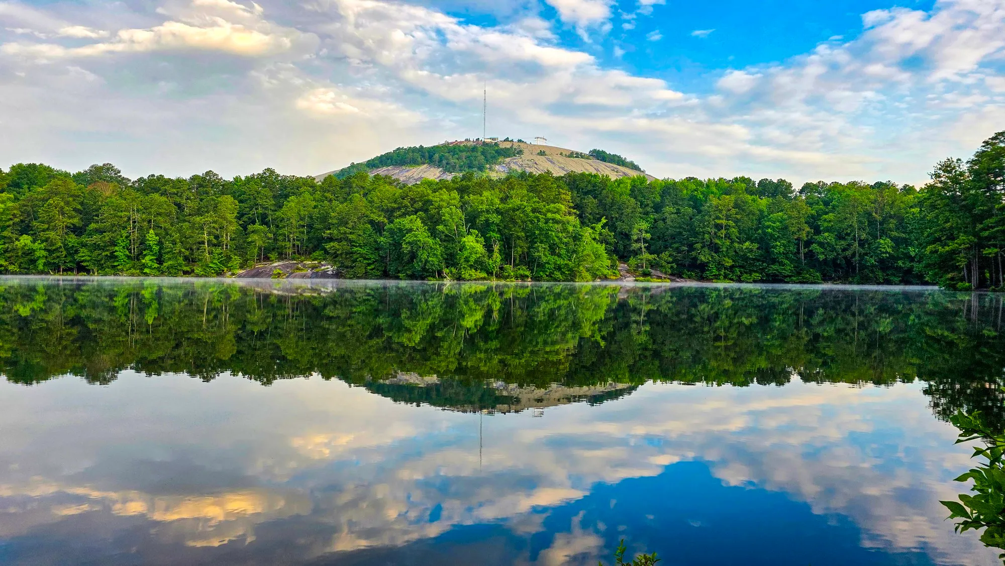 Stone Mountain and its tower above a wooded shore under blue sky and white cloud, the lake mirroring it