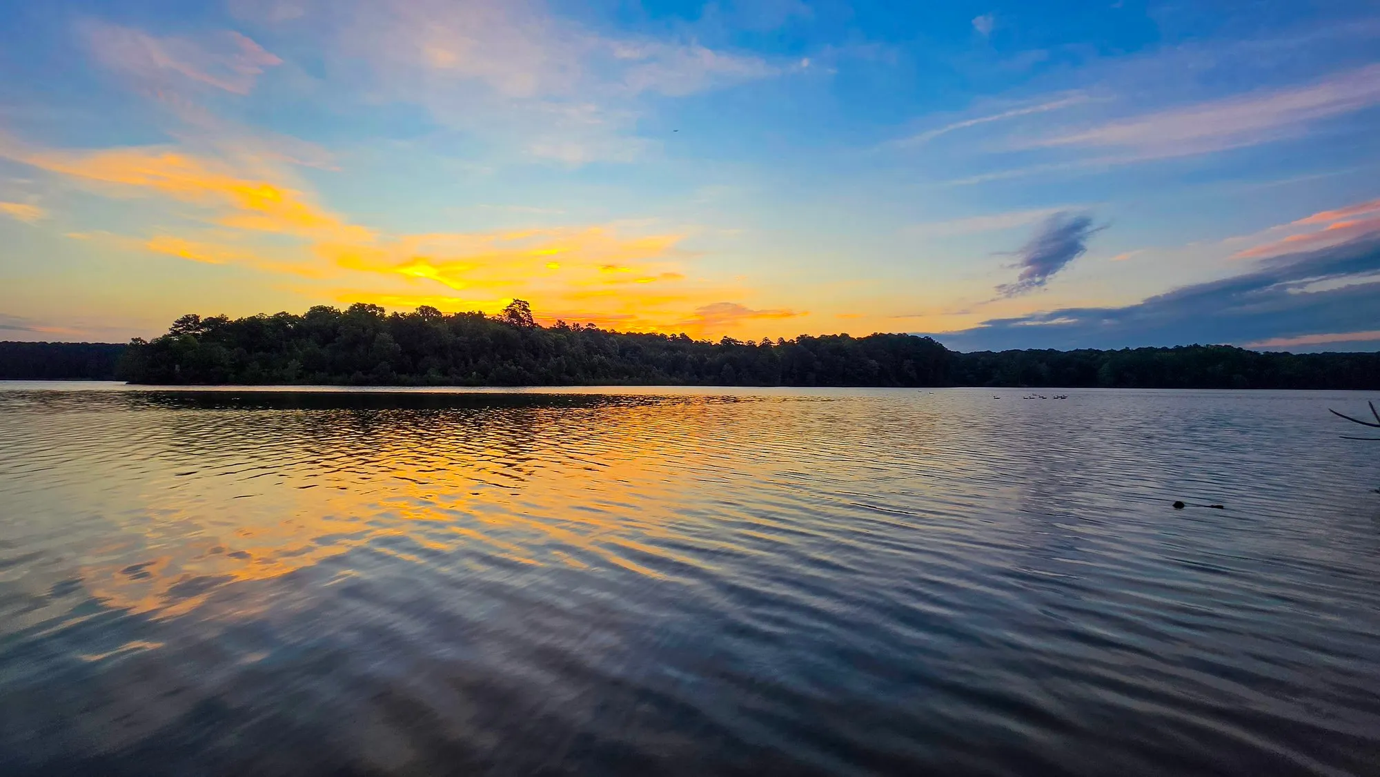 Gold light along the treeline of the far shore at sunrise on Stone Mountain Lake, a band of soft cloud above and light ripples on the water