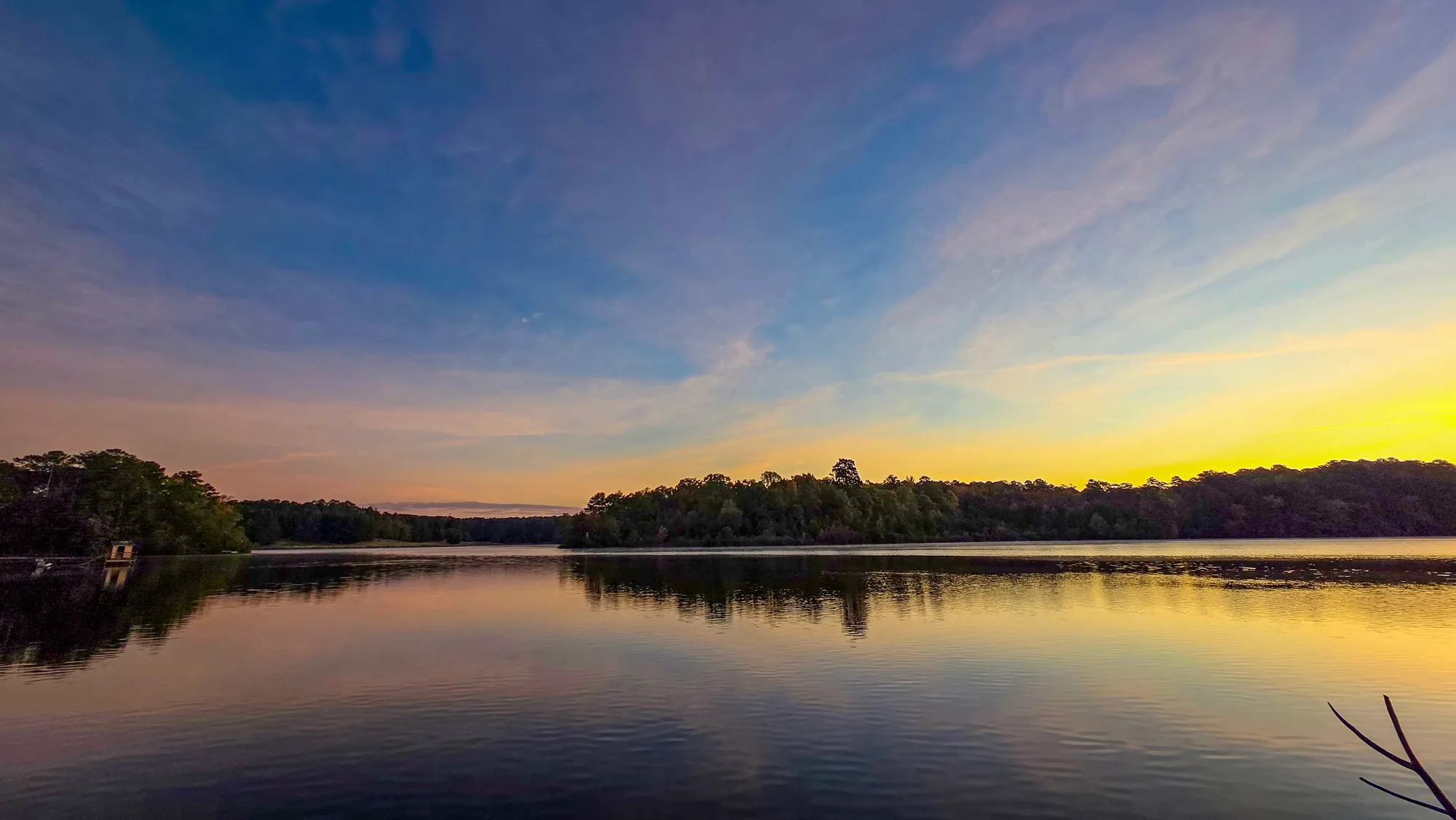 Thin high cloud streaked across a blue sky before sunrise, a band of yellow on the horizon, the lake calm