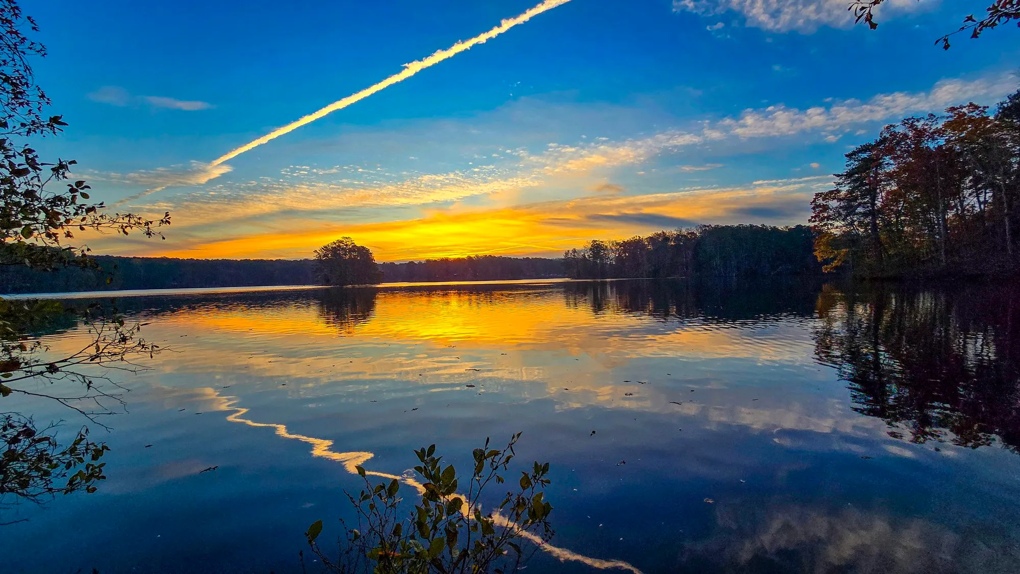 A bright contrail slanting across a blue and gold sky after sunrise, the lake still, autumn trees on the right