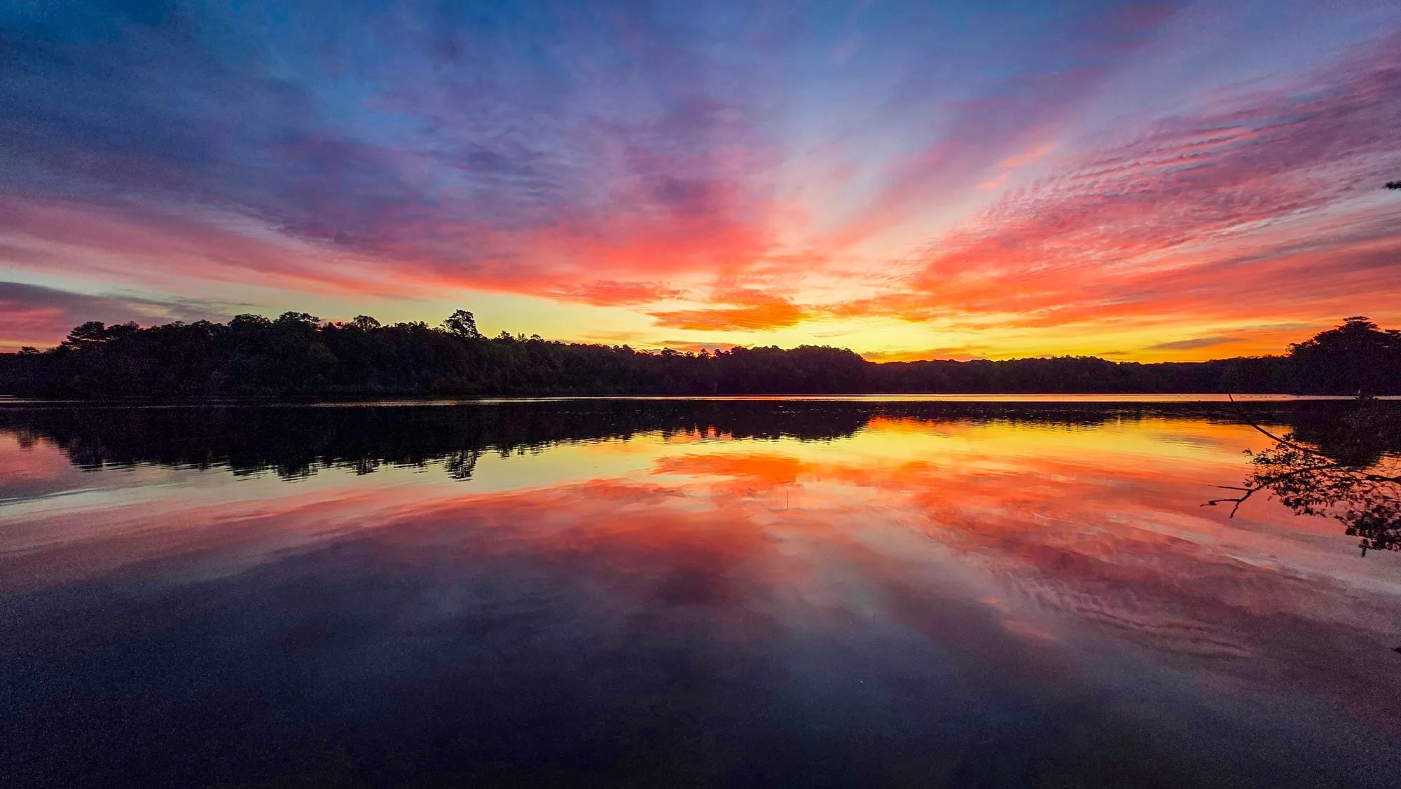 Red and pink cloud streaks radiating from the far shore before sunrise, the whole sky doubled in a still lake