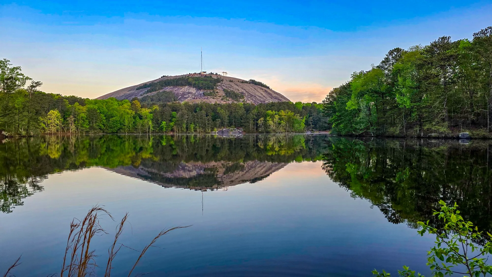 Stone Mountain mirrored in the still lake at sunrise, a faint pink glow beside the dome, fresh spring green on both shores