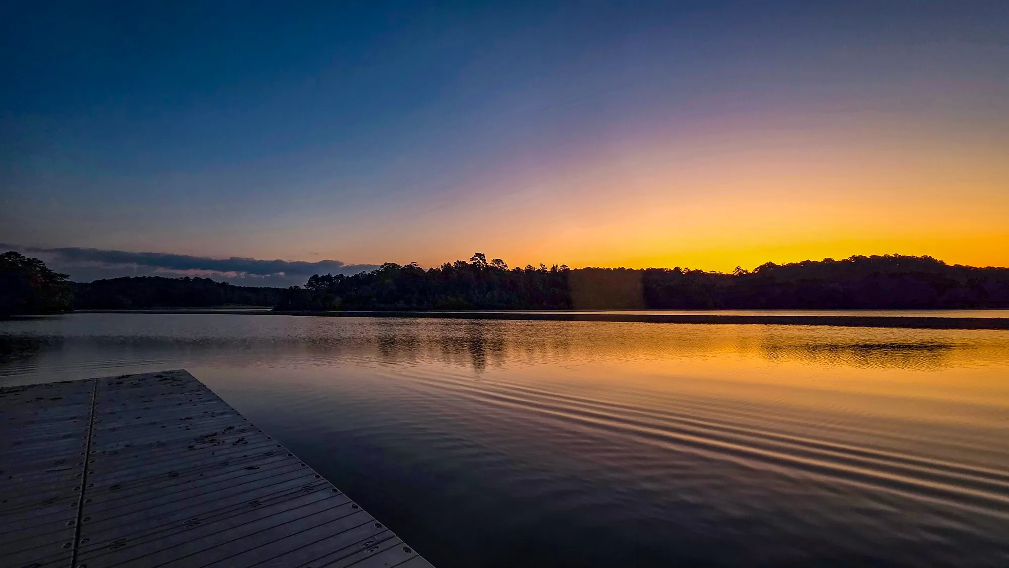 Gold and blue dawn over the far shore from a dock, ripples spreading across the lake