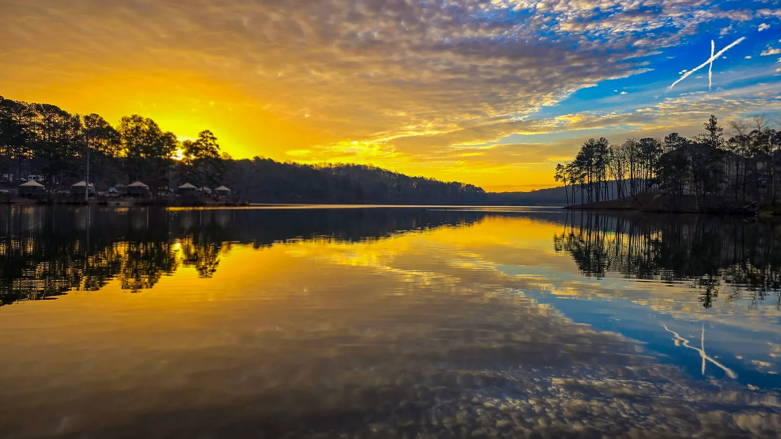 Yellow dawn sky with a contrail over the far shore, bare trees and cabins along it, the lake still and gold