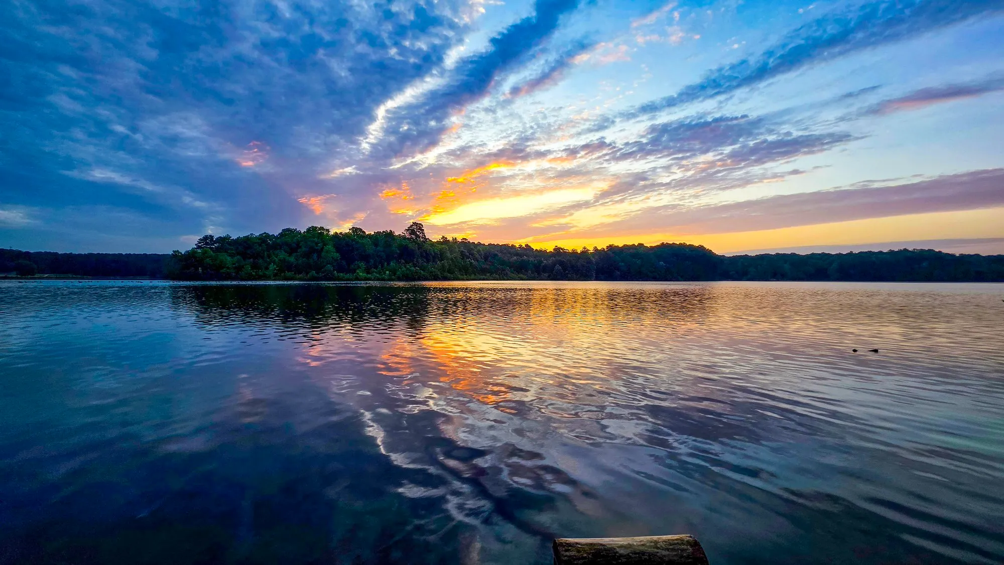 Rays of cloud fanning out from the sunrise over Stone Mountain Lake, blue above, orange at the treeline, ripples breaking the reflection