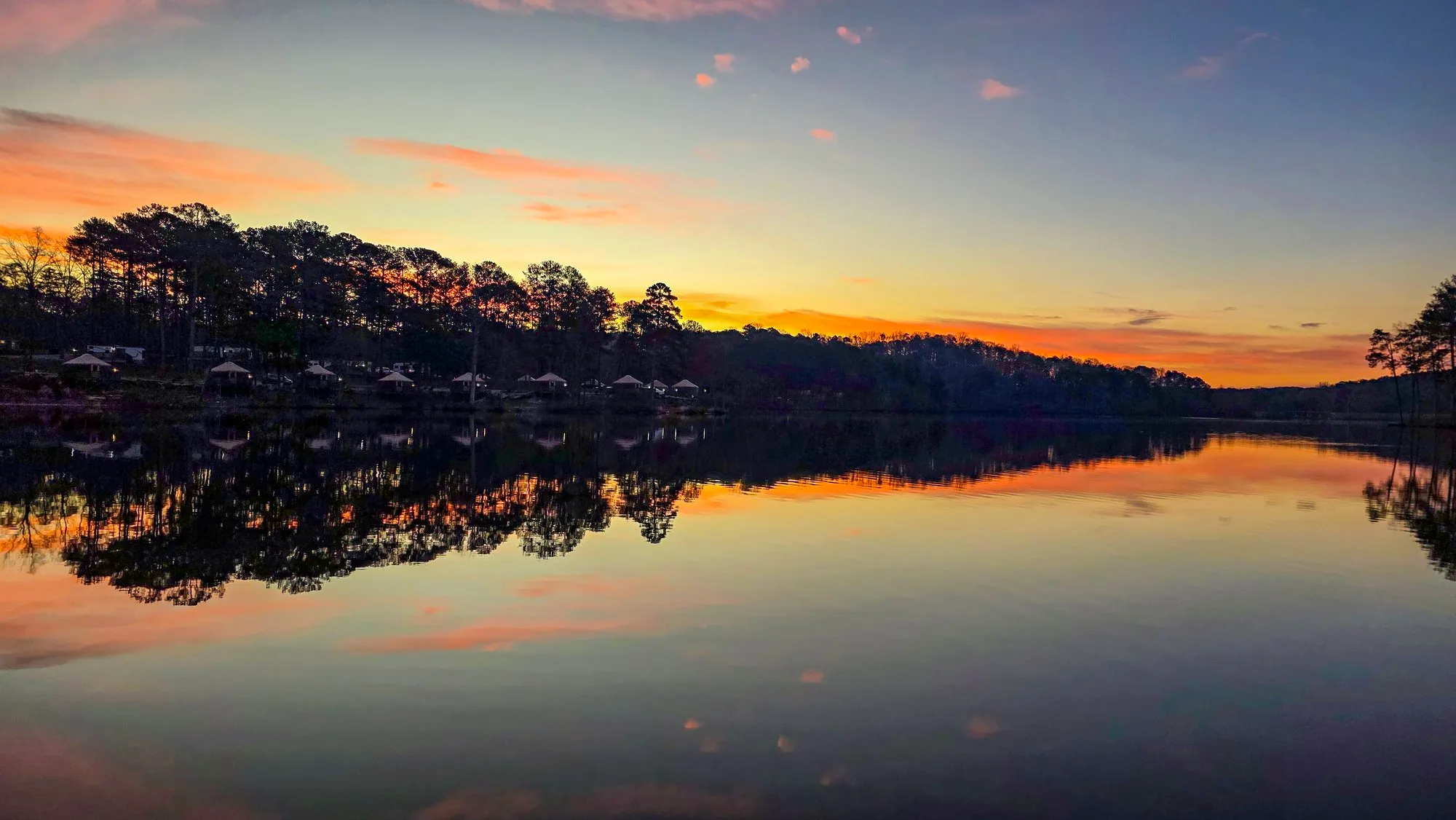An orange band of dawn under blue sky over the far shore, pink cloud above, the lake still and mirroring it