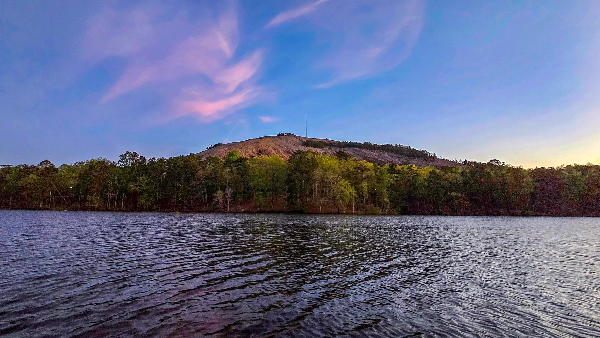 Pink wisps of cloud high over Stone Mountain across the lake, the summit mast against a blue sky, spring green along the shore