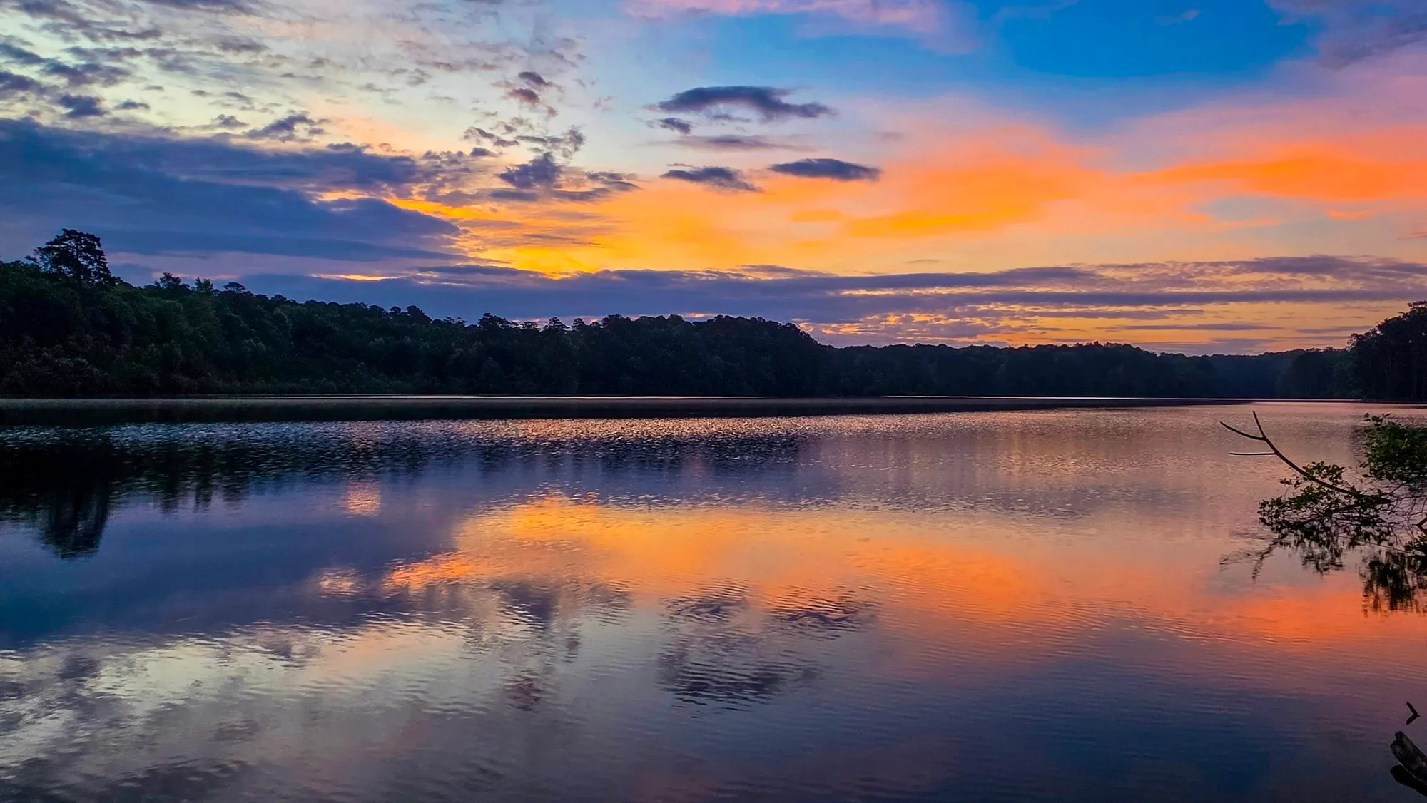 Orange cloud bands under a blue sky before sunrise, a dark tree line on the far shore, the lake mirroring the orange