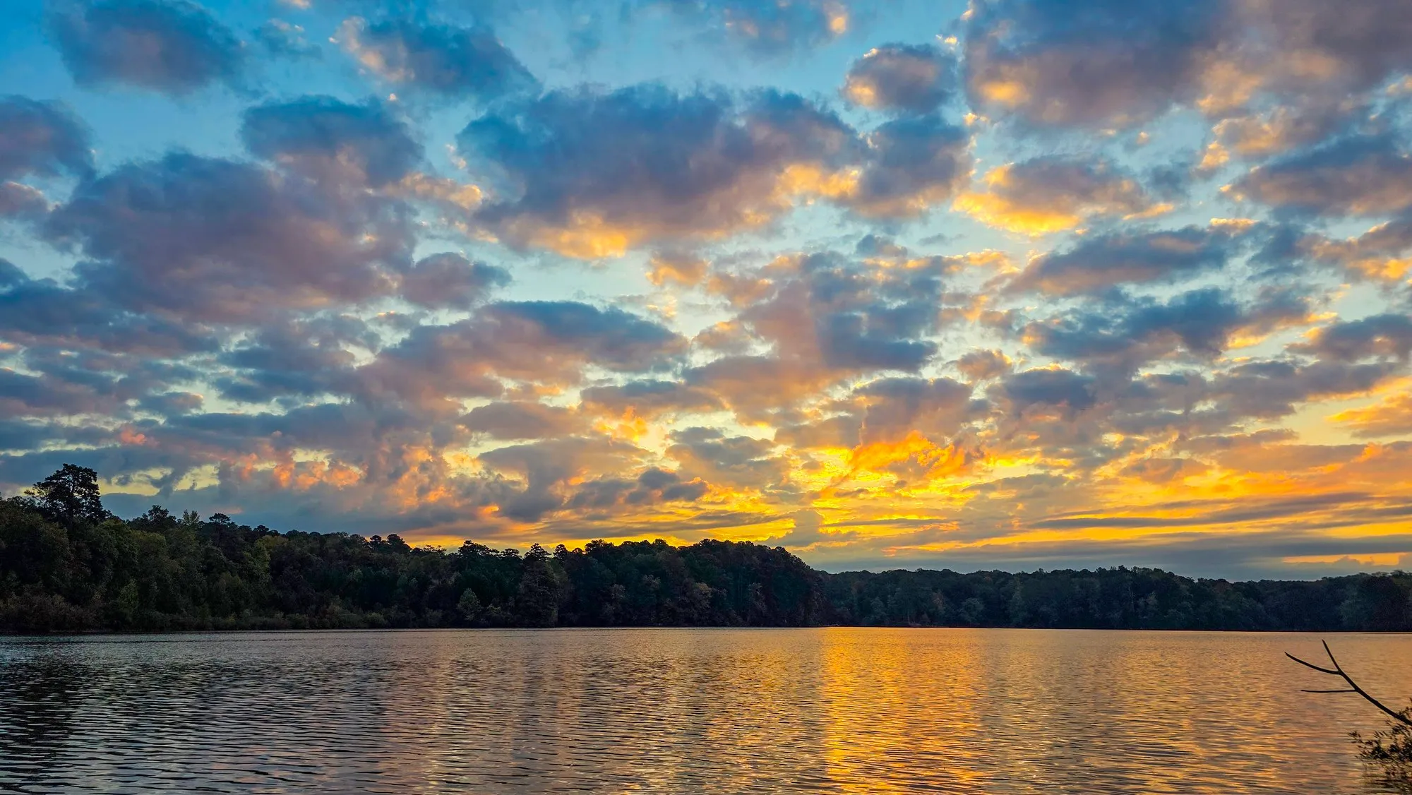 Rows of gray cloud with gold light breaking through after sunrise, the lake carrying the gold, trees on both shores