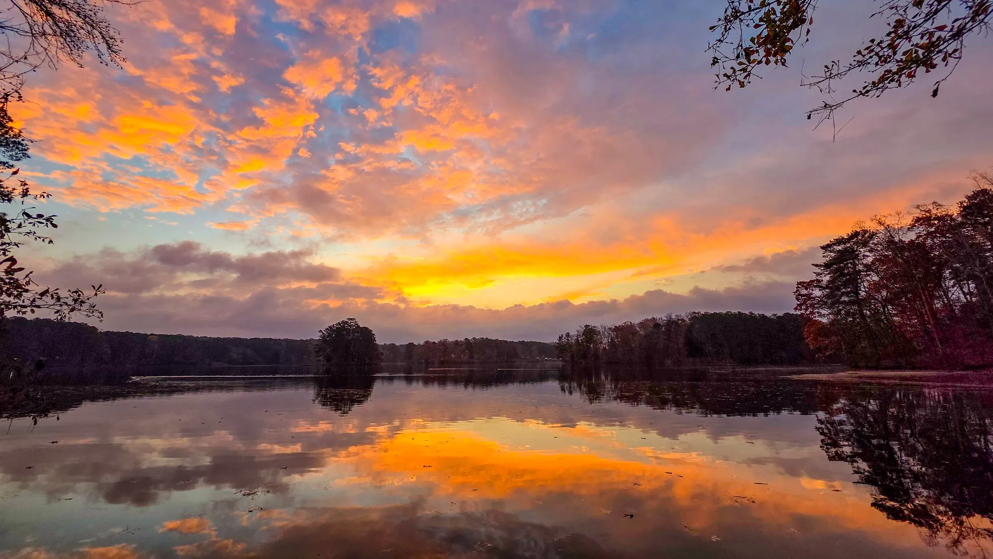 Pink and orange cloud fanning across the sky before sunrise, a few autumn leaves in the top right, the lake mirroring the color