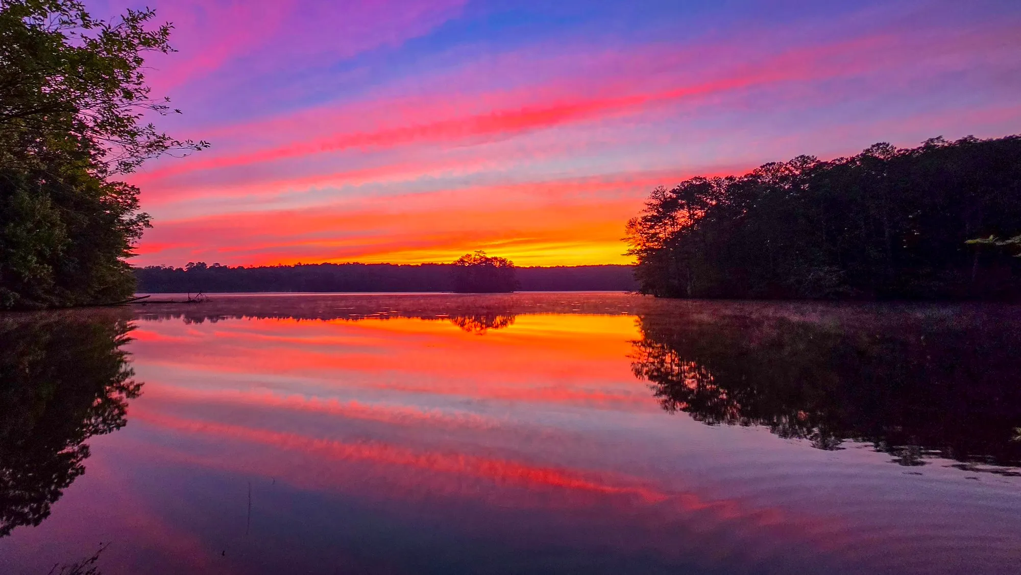 Pink and red cloud streaks across the whole sky before sunrise, mirrored in the lake, the far shore a dark line