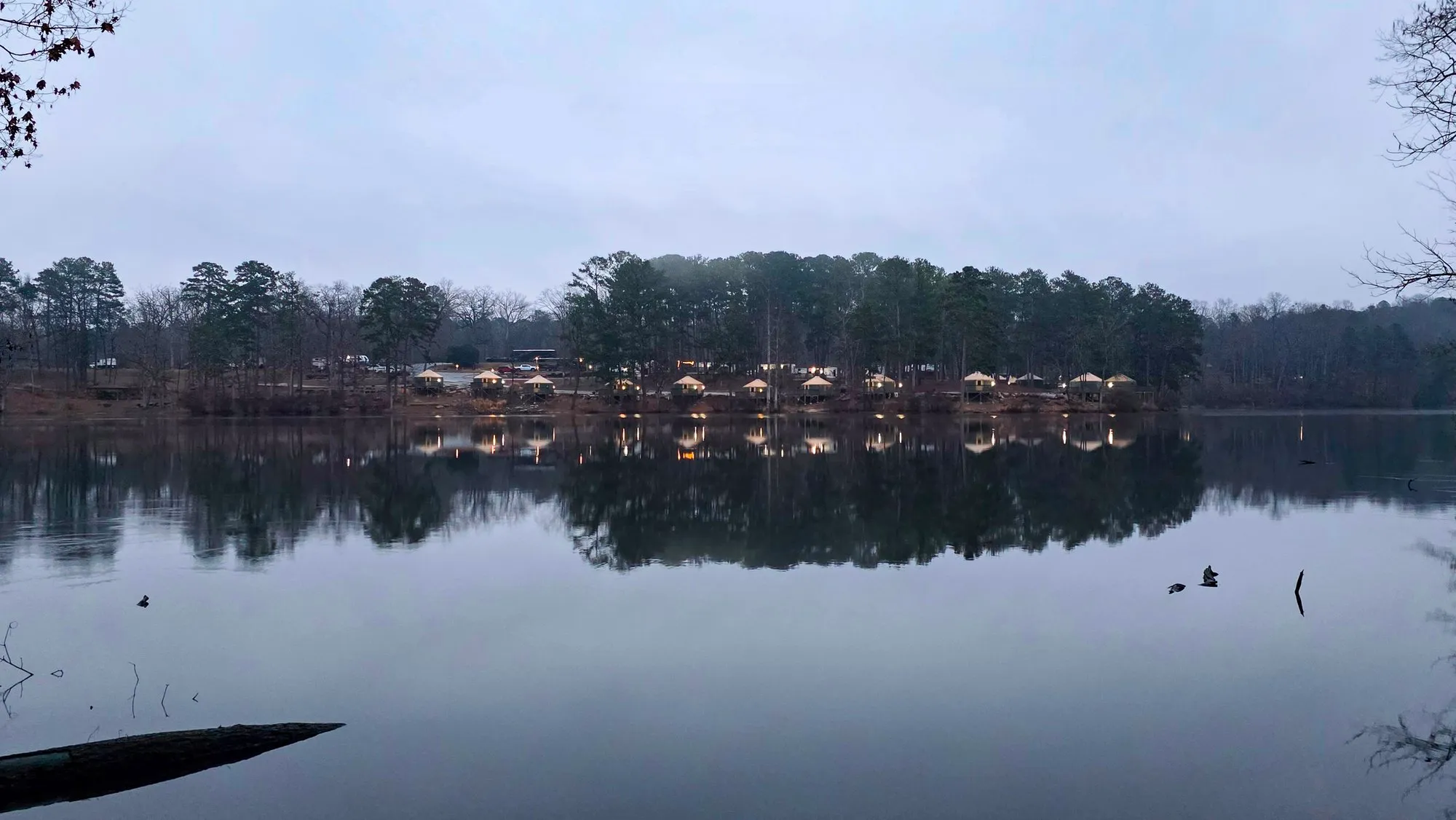 A still gray winter morning on the lake, low cloud over the far trees and their lit buildings mirrored in flat water, a fallen branch at the shore