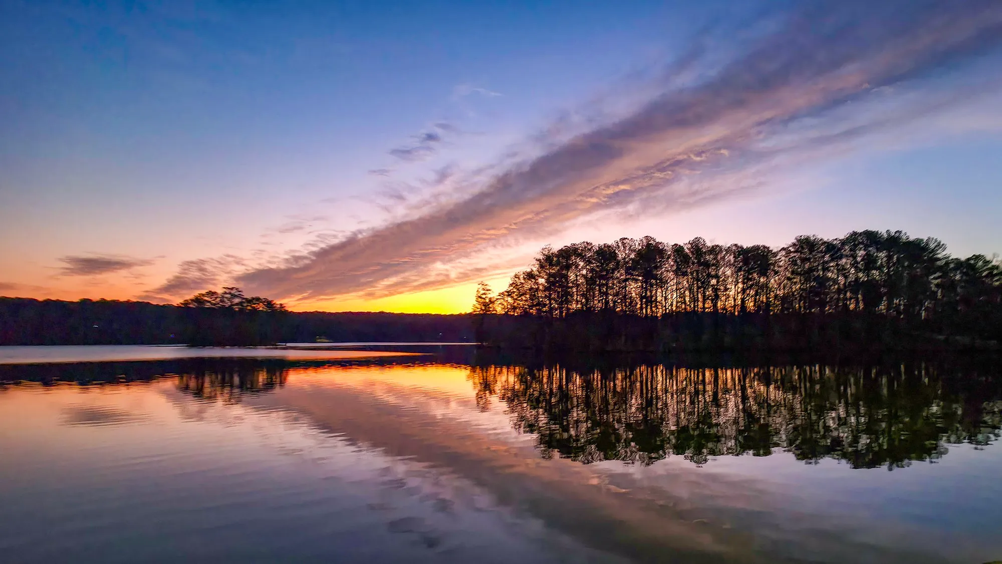 A dark band of cloud crossing a gold dawn sky, pines on the far point, everything doubled in the still lake