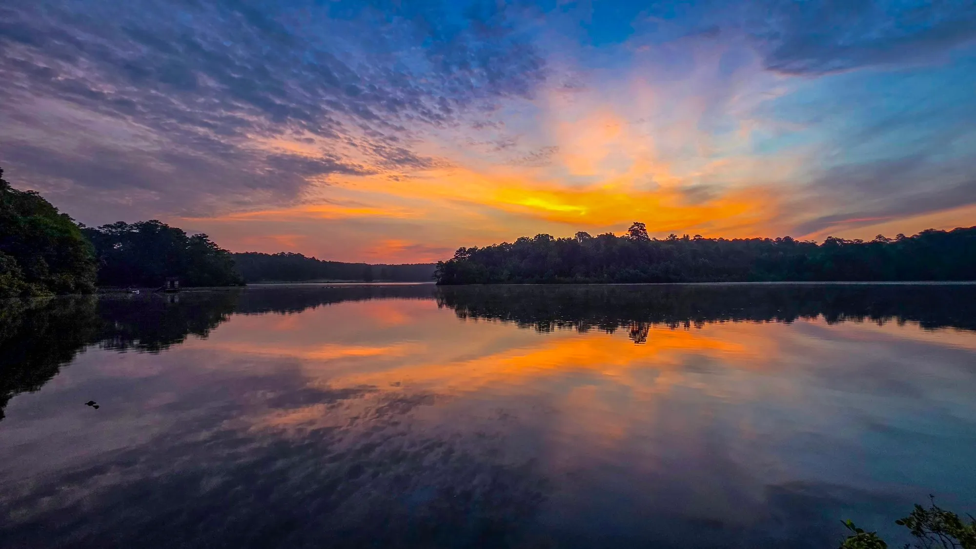 Pink and gold cloud radiating from behind the far shore of Stone Mountain Lake before sunrise, mirrored in calm water