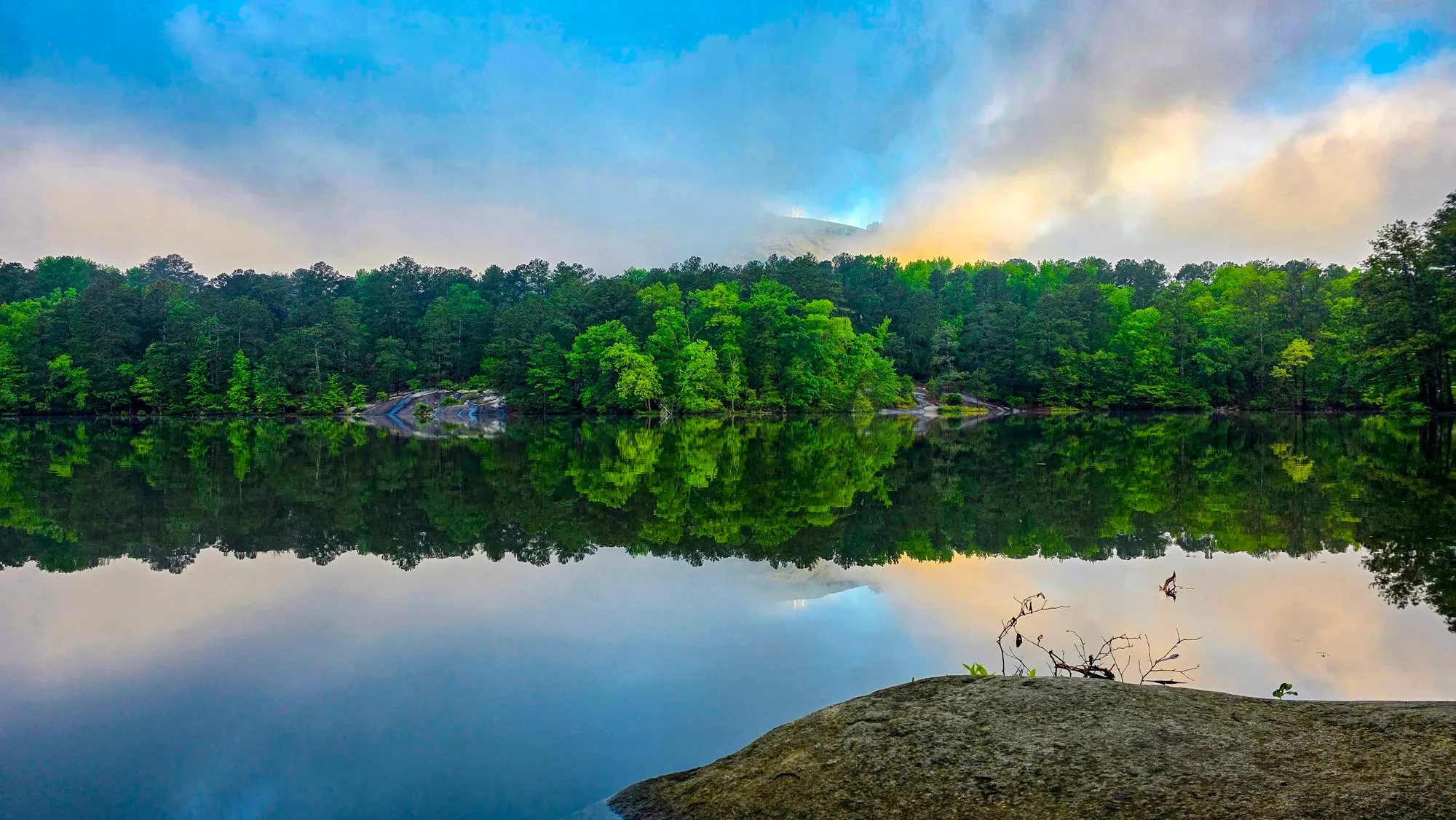 Green shoreline and a rock outcrop reflected in a still lake under broken cloud and blue sky after sunrise