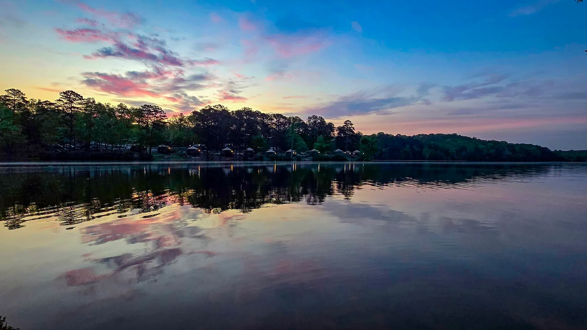 Pink and violet cloud streaks across a blue sky before sunrise, mirrored in the calm lake, the campground lights along the far shore
