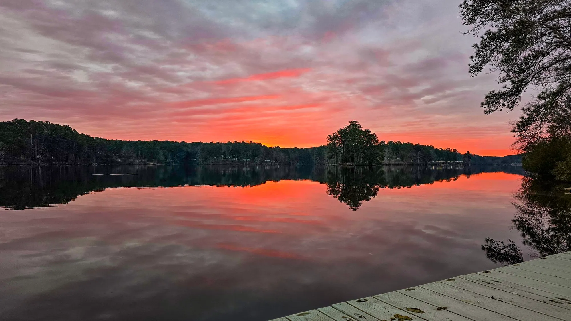 Red and pink cloud across the whole sky at sunrise, mirrored in the still lake, a wooden dock in the right corner and a pine island in silhouette