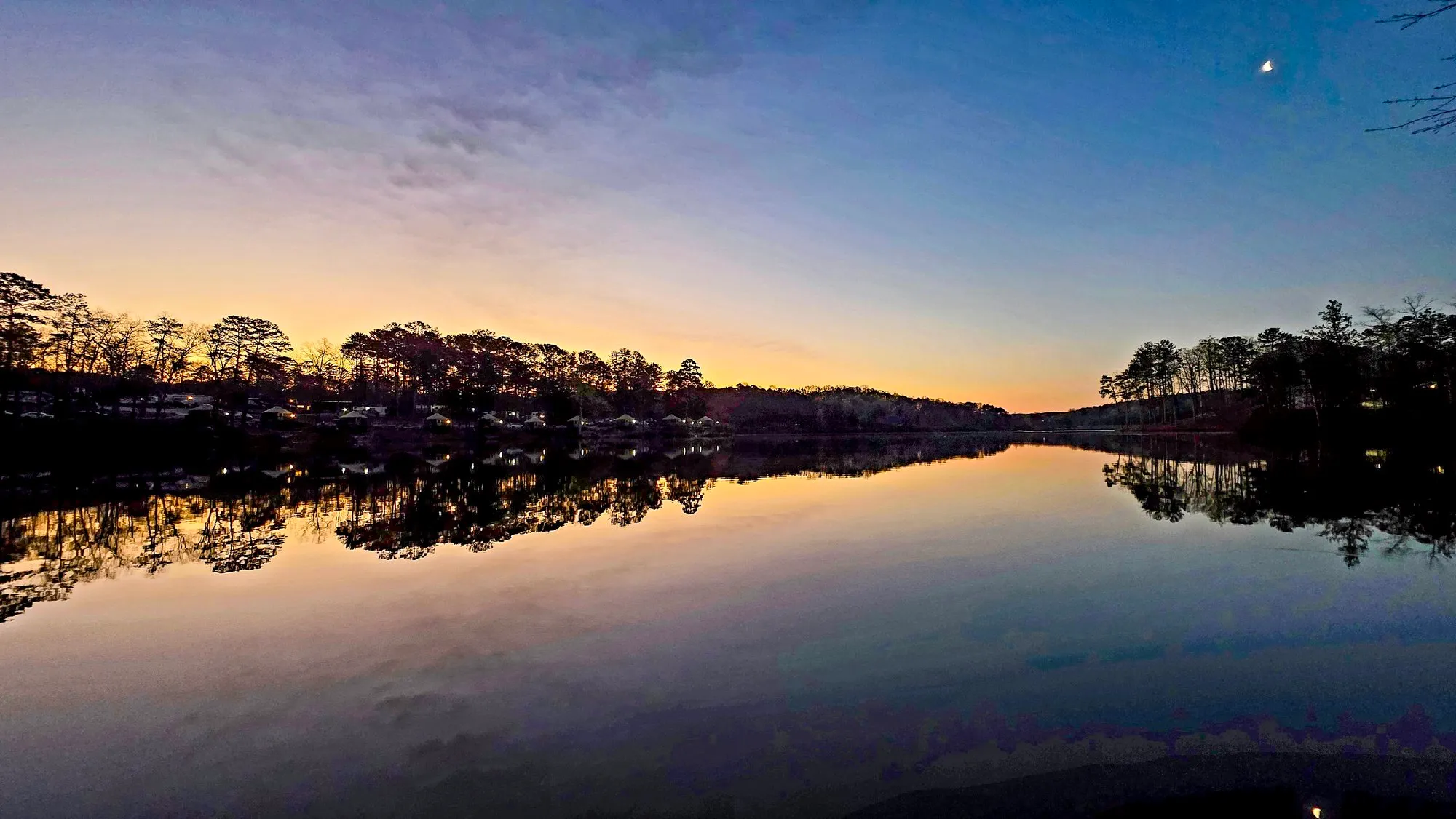 Blue and gold dawn with a sliver of moon high in the sky, the far shore dark, the water still