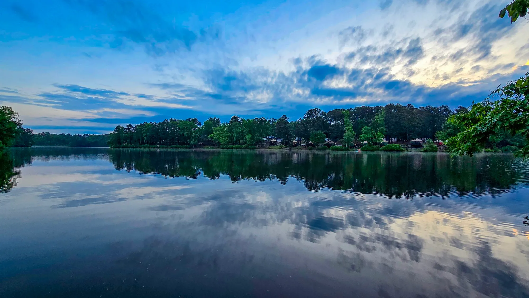 Blue sky with streaks of white and gray cloud after sunrise over the far shore, mirrored in a still lake, leaves hanging in at the right edge