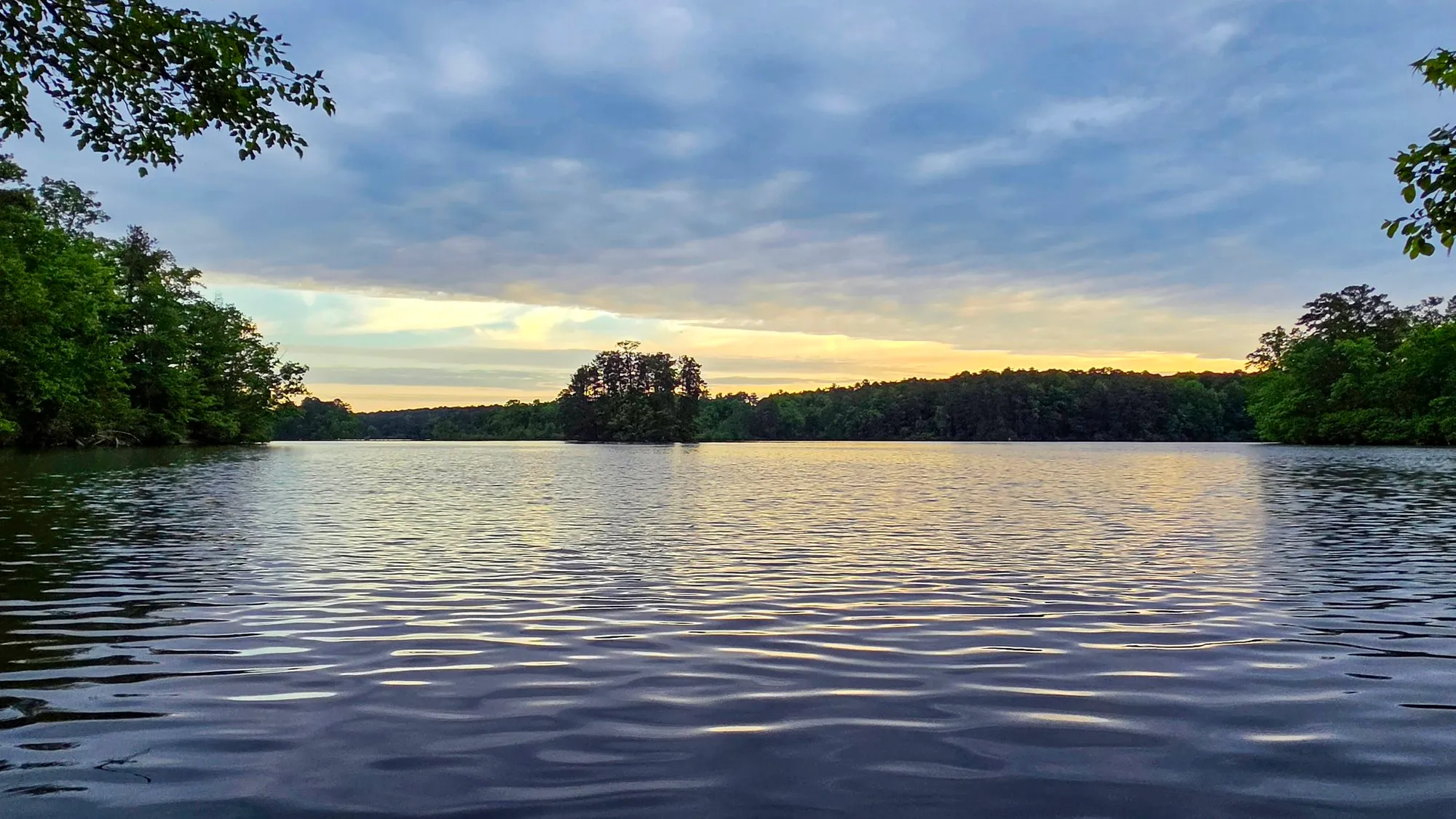 Blue and gray cloud over the lake after sunrise with a band of yellow light on the horizon, a wooded point in the middle distance, branches framing the top