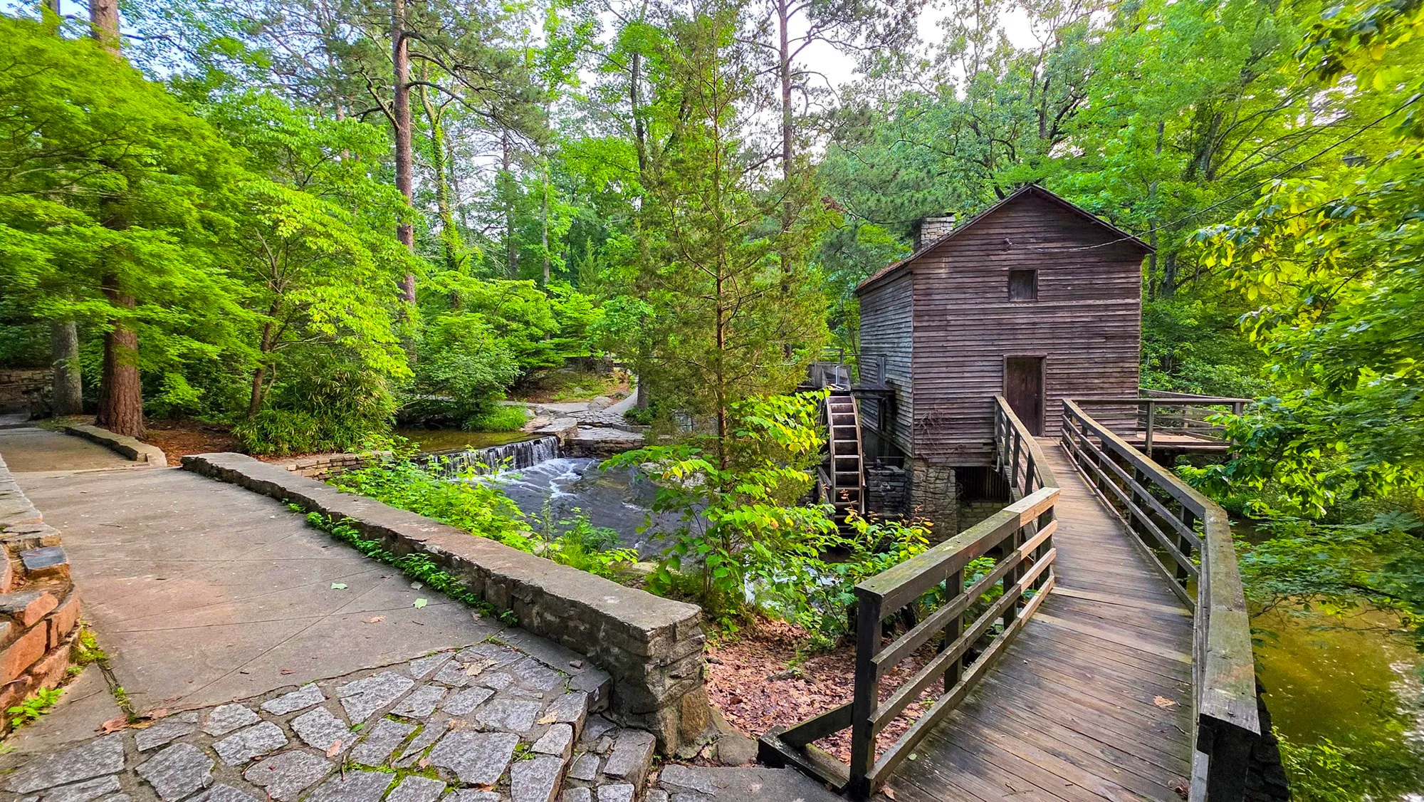 The Grist Mill above its cascade, the wooden footbridge crossing to it on the right and the stone-paved path on the left, everything green in the last week of May