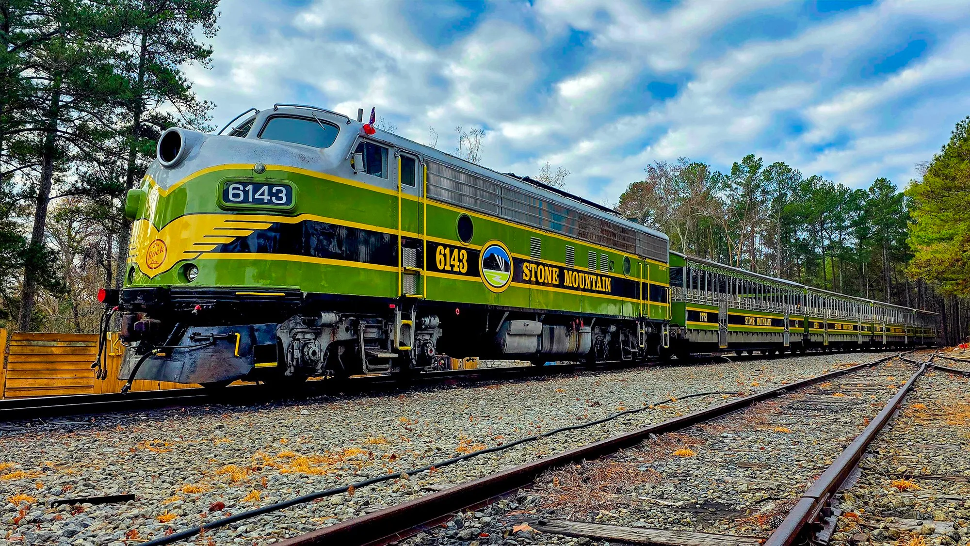 The park's green and yellow locomotive 6143 at the head of its open cars, stopped on the tracks under a mackerel sky, the rails curving away past the pines