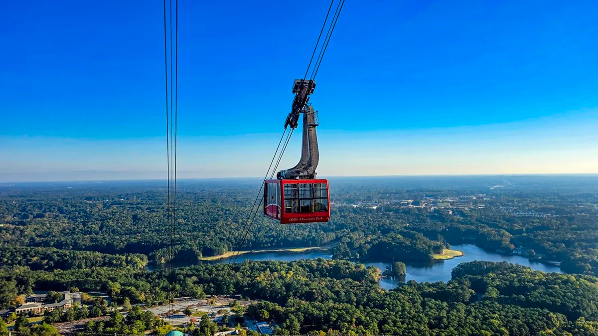 A red Summit Skyride car on its cables above the forest, seen from high on the mountain, with the lake winding through the trees below and wooded land running out to a hazy horizon under a clear blue sky