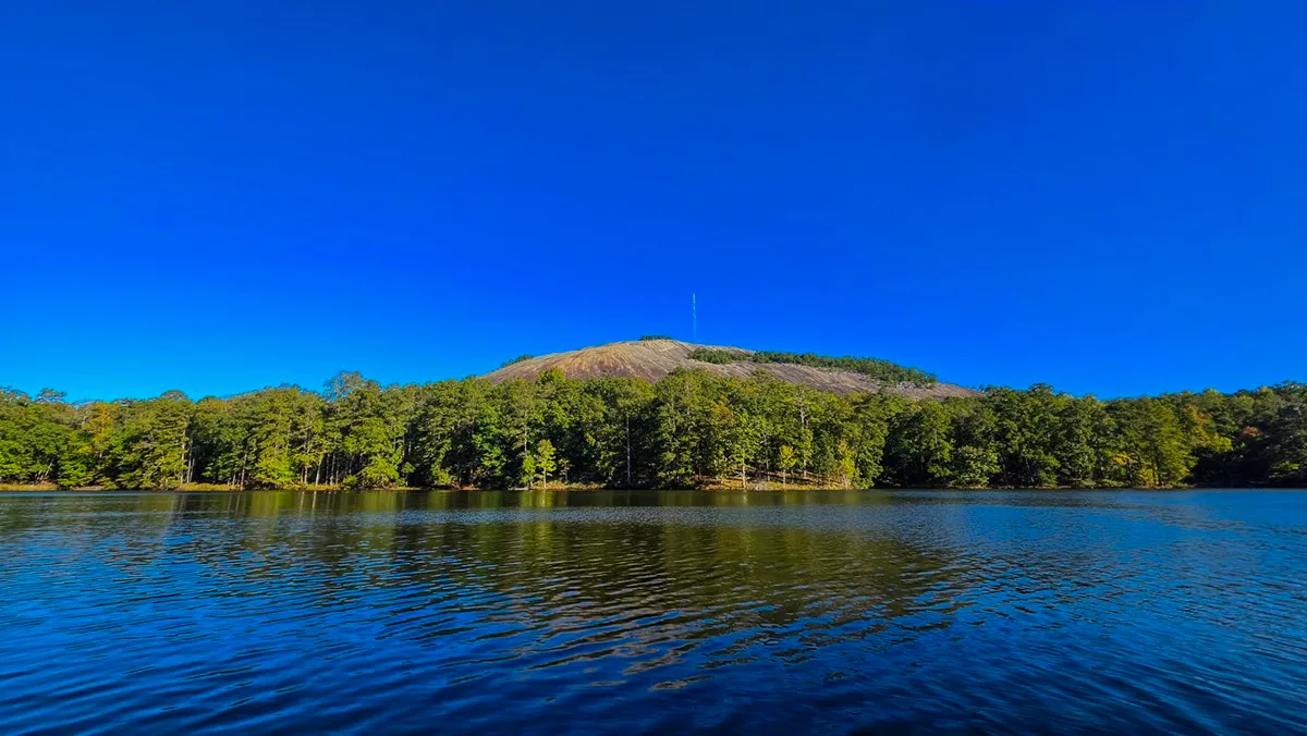 Stone Mountain's bare granite dome and its summit mast above green trees across Venable Lake, under a clear deep blue sky, the water rippled by the wind