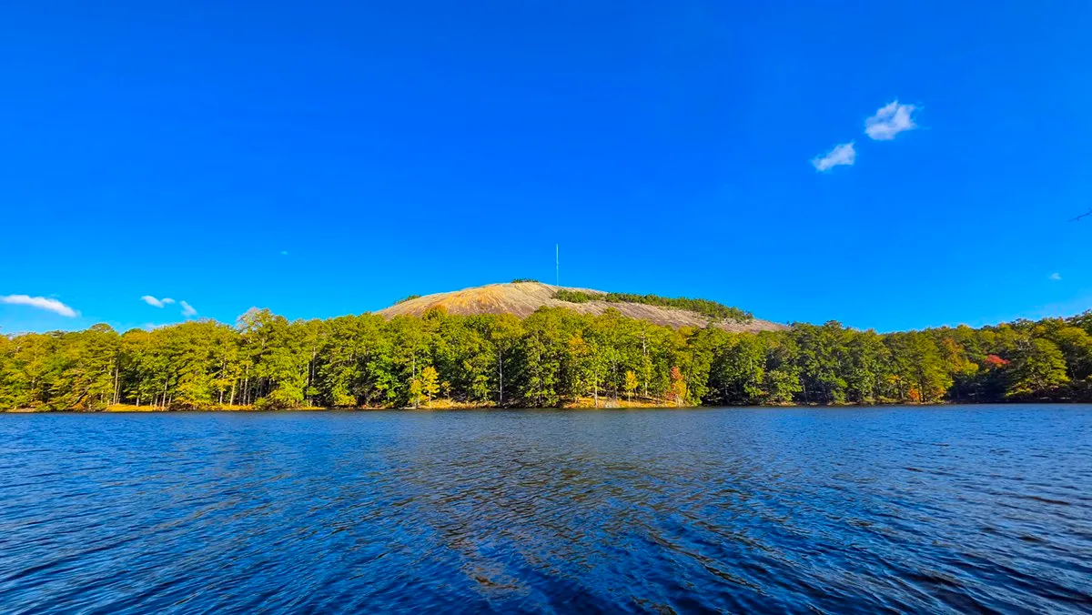 Stone Mountain's bare granite dome and its summit mast above a line of green trees across Venable Lake, a few red and orange leaves among them and a few small white clouds in a deep blue sky